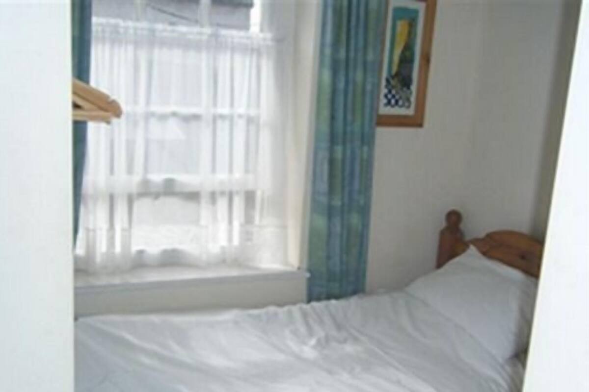 Ein kleines Zimmer mit einem Einzelbett, einem Fenster, einem Vorhang und einem gerahmten Bild an der Wand.