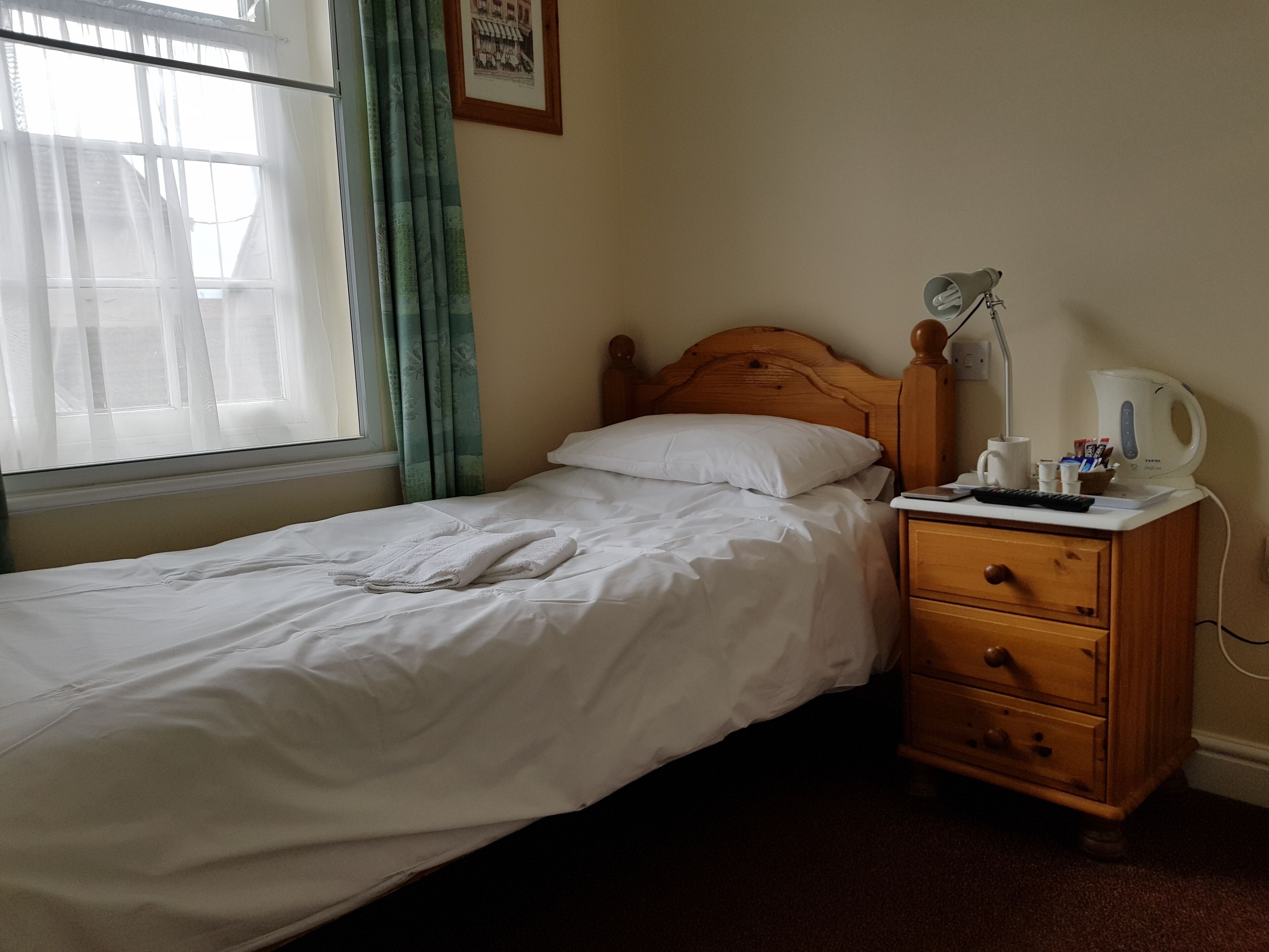 A single bed with white linens, a wooden nightstand with a lamp, a kettle, and a cup.