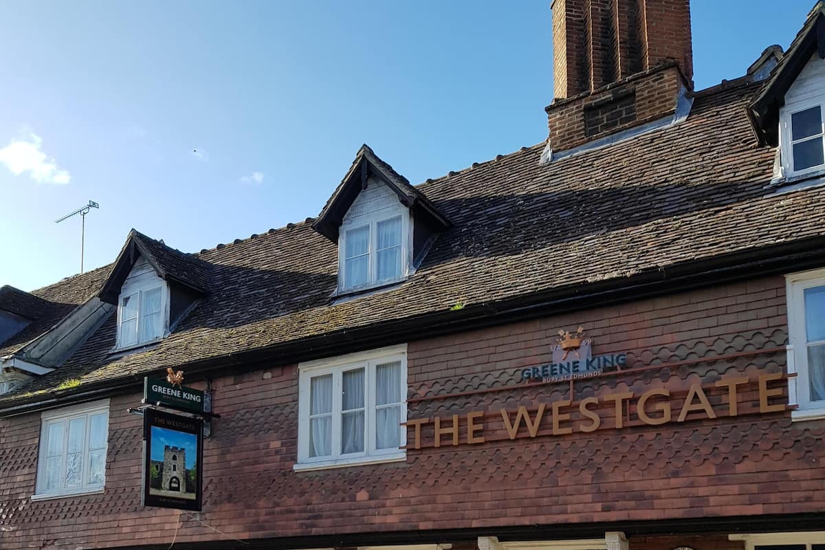 Ein traditionelles Backsteingebäude mit einem Schild, auf dem "The Westgate" und "Greene King" stehen, was auf ein Pub hinweist. Schilder für Billard, Darts und Fußball deuten auf Freizeitaktivitäten hin.