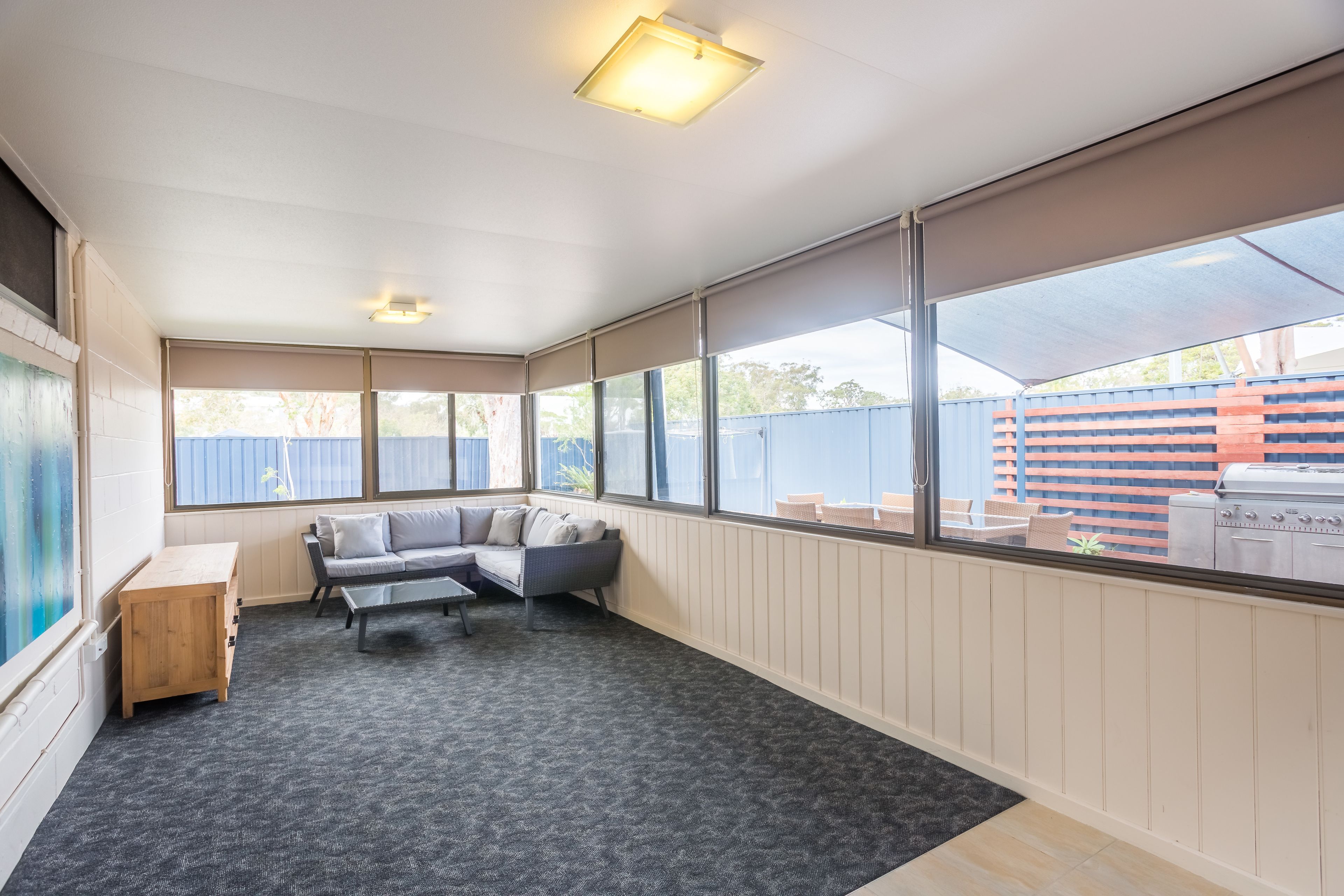 A covered patio area with a grey sectional sofa, a wooden table, and a large window overlooking a backyard with a barbecue grill.
