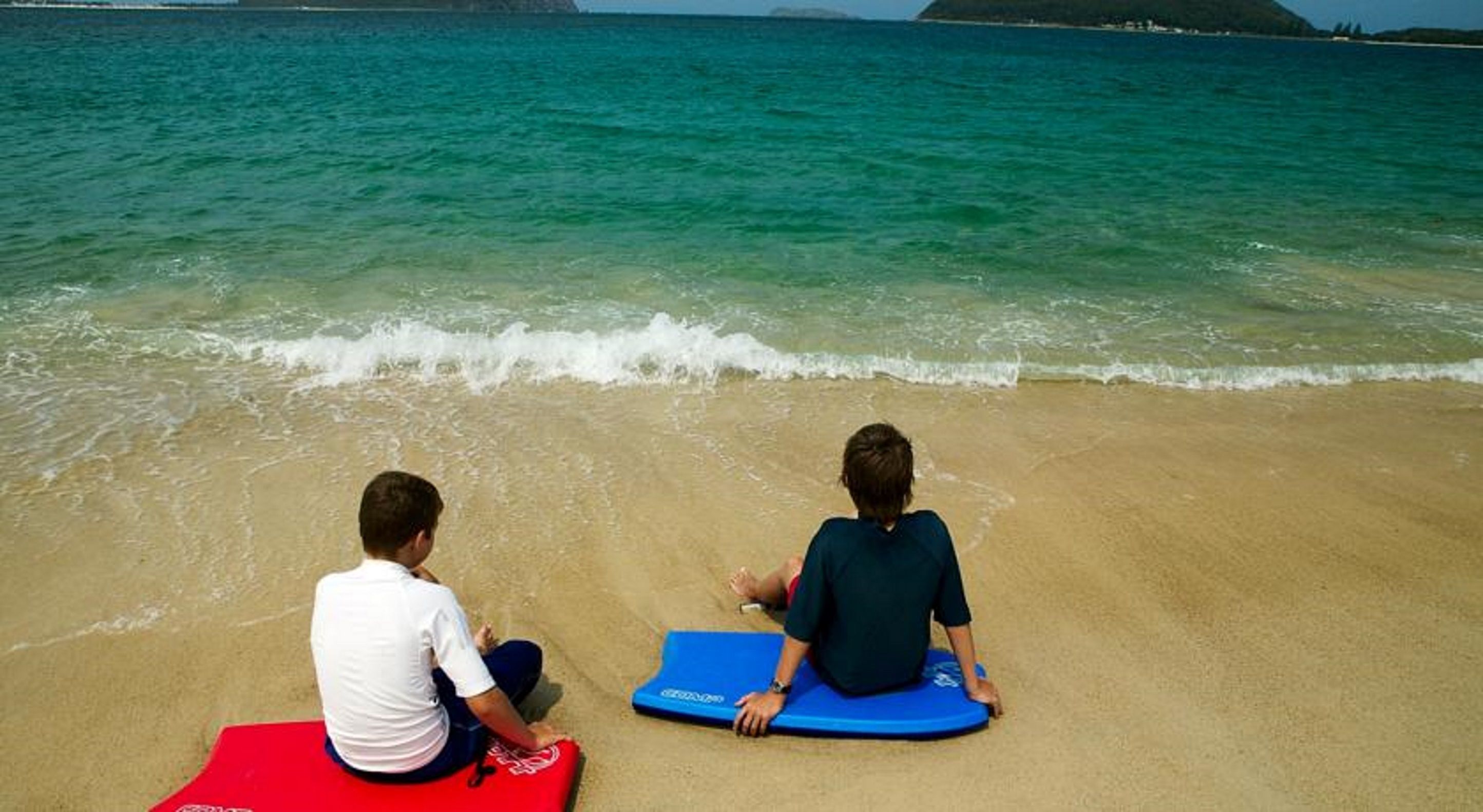 Zwei Personen sitzen auf Matten am Strand, mit klarem Wasser und einer entfernten Insel im Hintergrund.