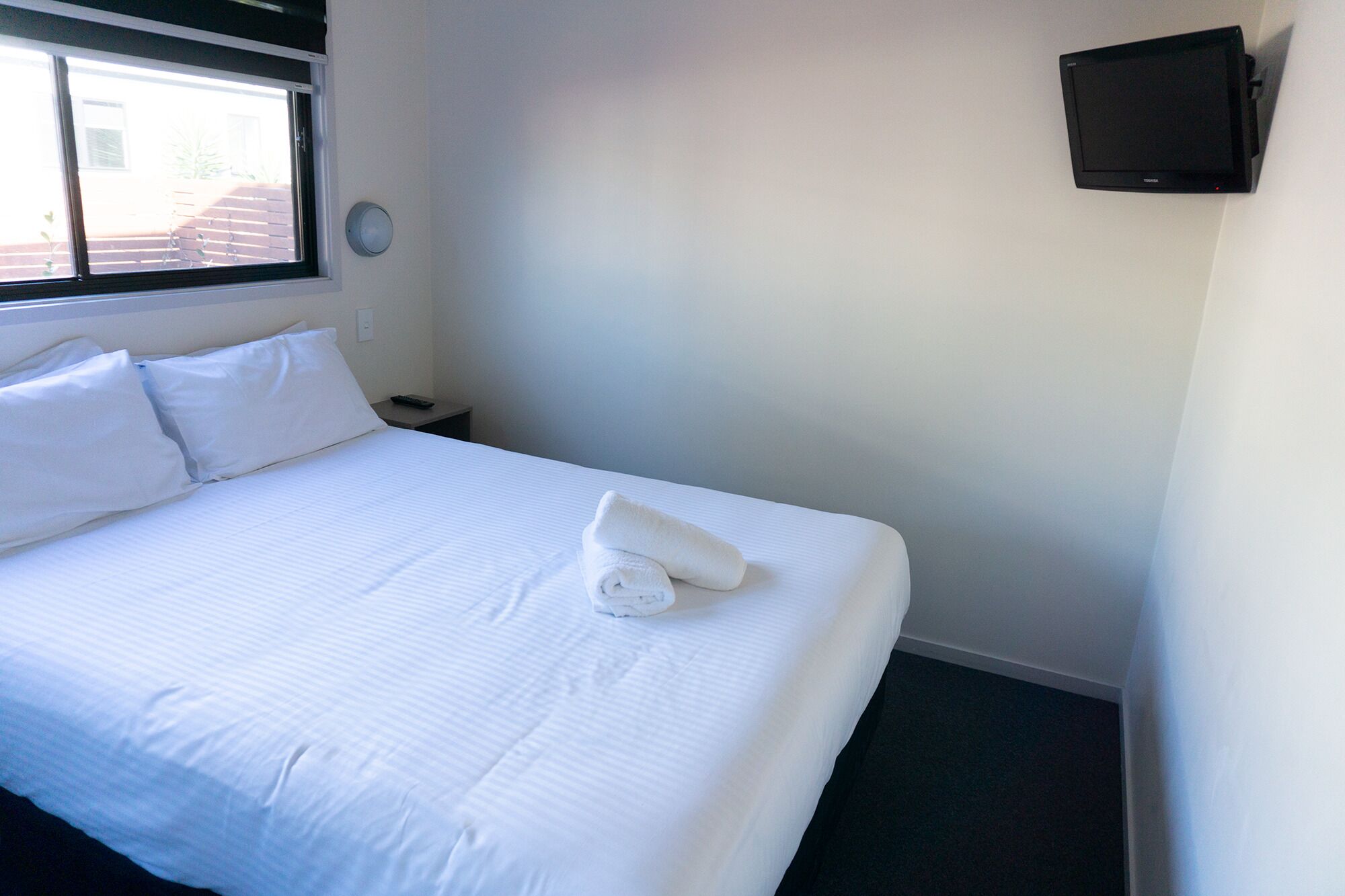 A hotel room with a neatly made bed, a bedside table with a remote control, a wall-mounted TV, and a window with blinds.