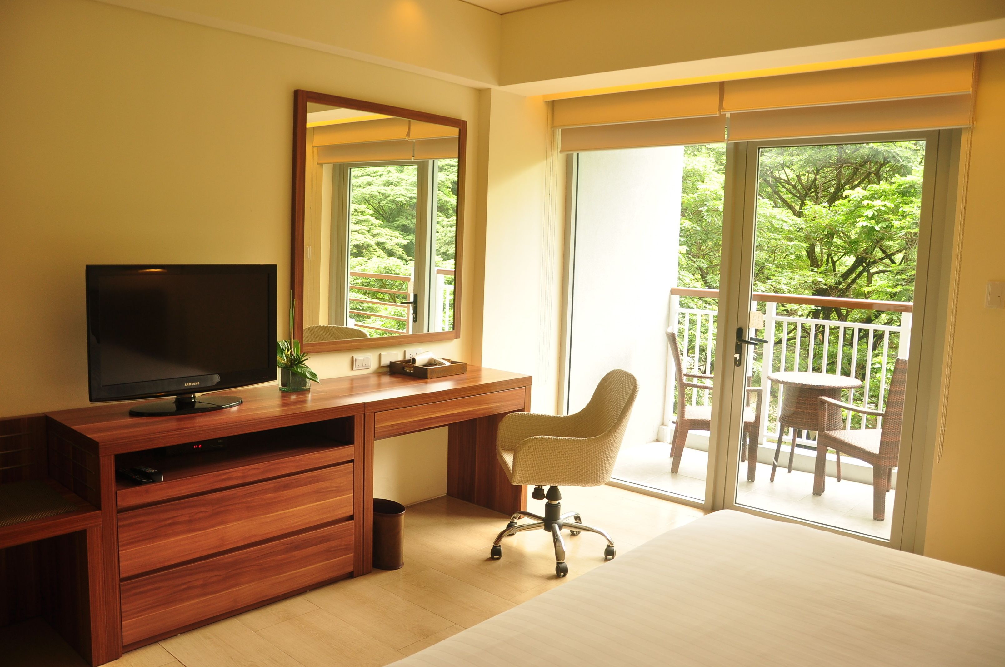 A bedroom with a wooden desk, a television, a chair, and a balcony with outdoor furniture.