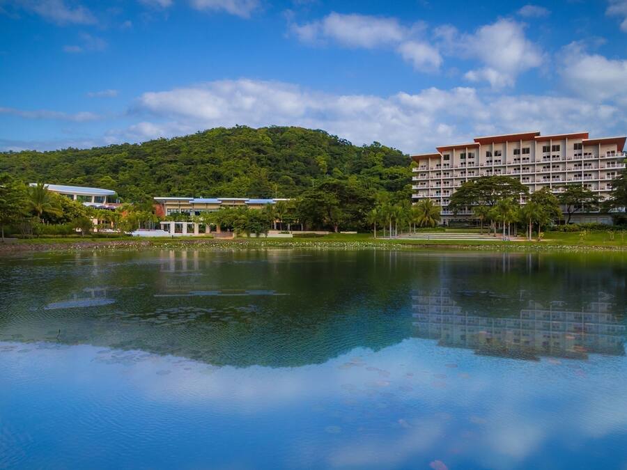 Un hotel de varios pisos, con un extenso cuerpo de agua al frente y una colina verde y exuberante al fondo.