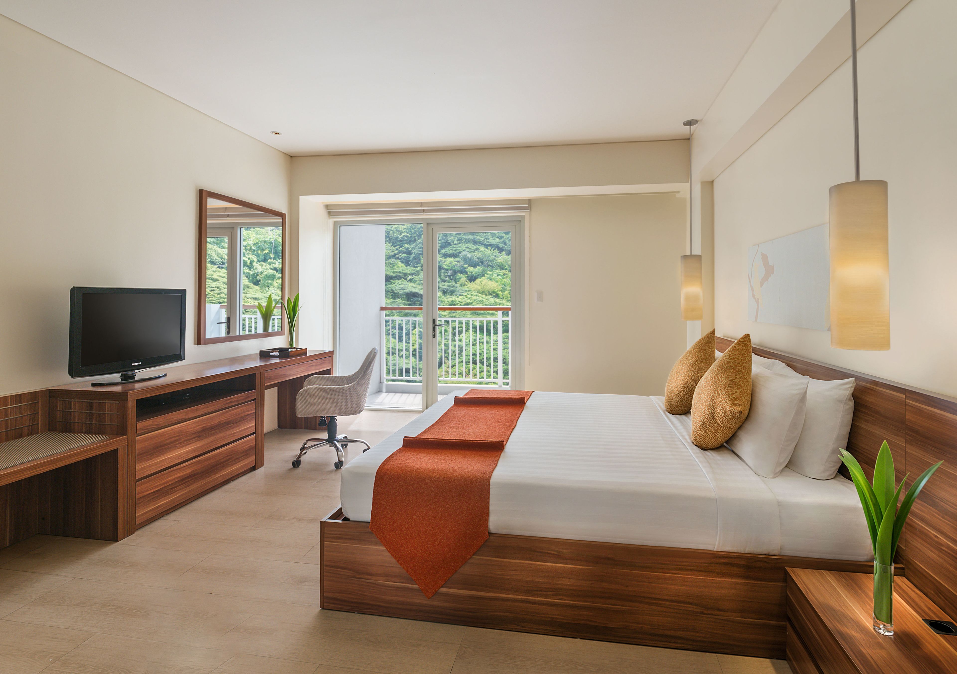 A modern hotel room with a wooden desk, a TV, a comfortable bed, a balcony with a view of greenery, and a large window.