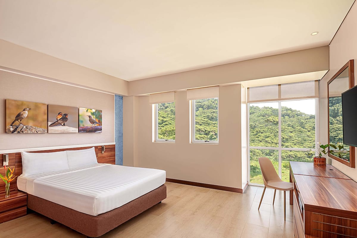 Ein modernes Hotelzimmer mit einem großen Bett, einem Schreibtisch und Blick auf üppiges Grün durch große Fenster.