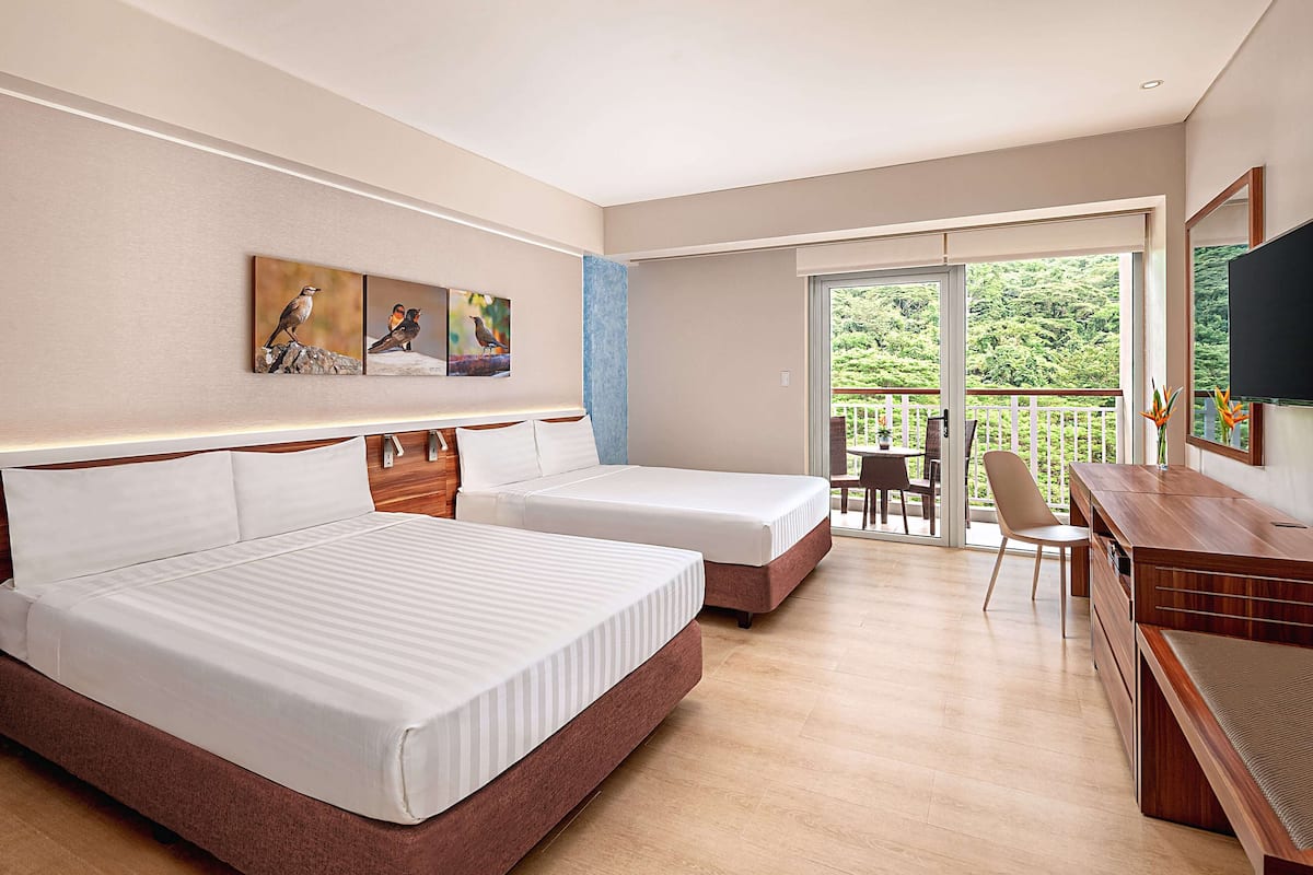 Ein Hotelzimmer mit zwei Betten, einem Schreibtisch, einem Stuhl, einem Fernseher und einem Balkon mit Blick auf Grünflächen.