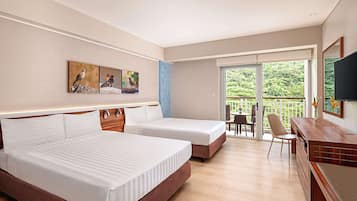 Habitación de hotel con dos camas, un escritorio, una silla, un televisor y un balcón con vistas a zonas verdes.