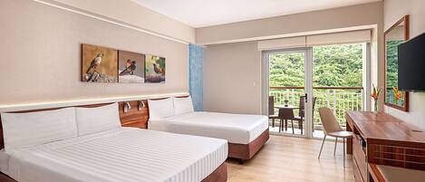 A hotel room with two beds, a desk, a chair, a TV, and a balcony with a view of greenery.