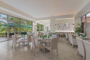 A spacious indoor dining area with round tables and white wicker chairs, large windows with palm trees visible outside, and a modern wall art piece. - Pico Sands Hotel (Nasugbu)