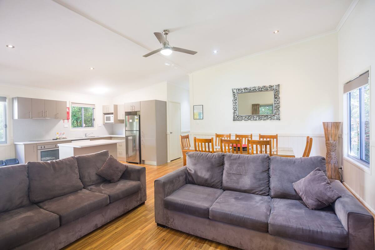 A modern living room with two grey sofas, a dining area with wooden chairs, a kitchen with white cabinets, and a ceiling fan.