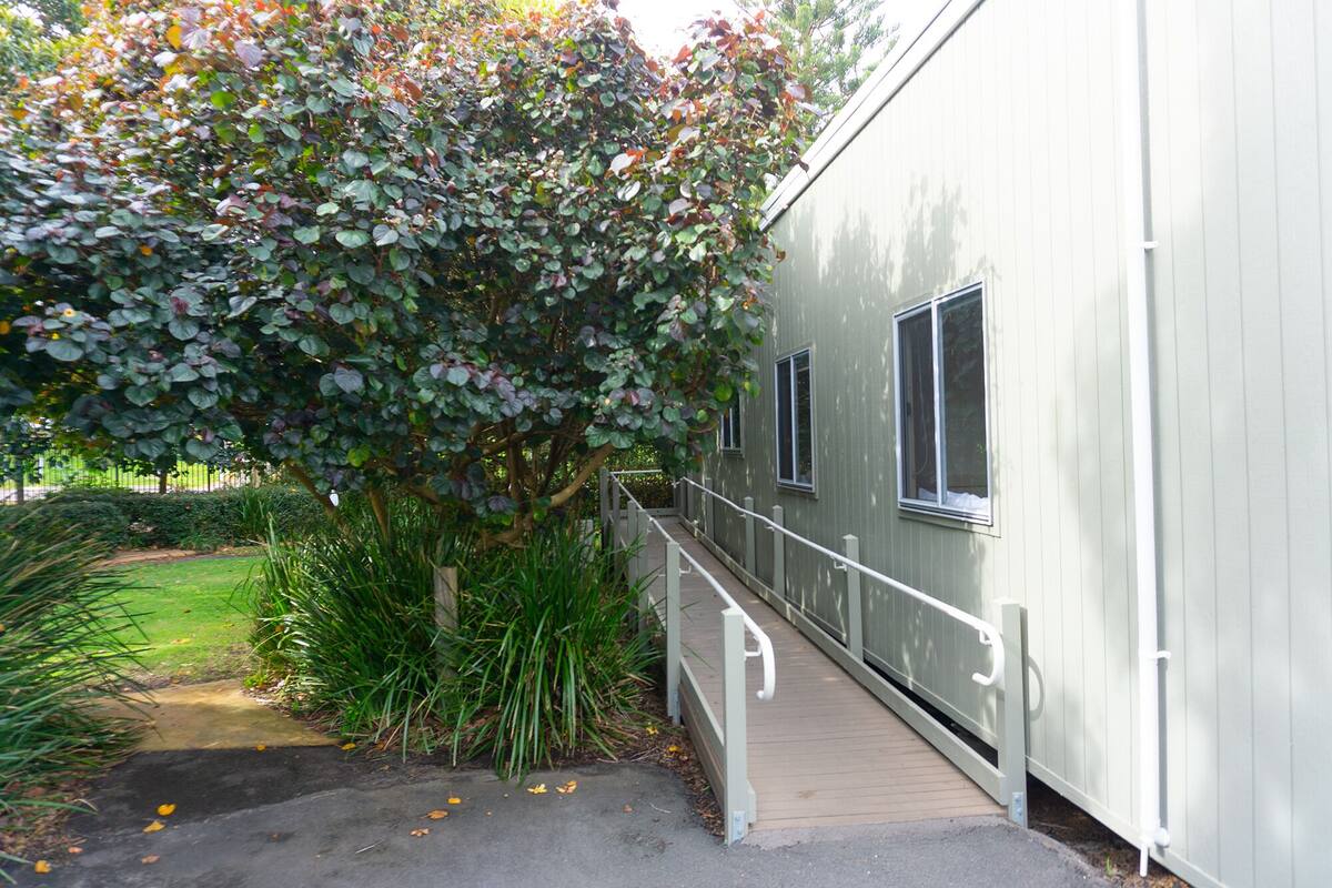 A building with a wooden deck leading to a gated entrance, surrounded by greenery and a small garden area.
