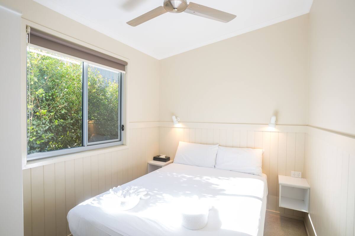 A bedroom with a bed, bedside tables, a nightstand, a ceiling fan, and a window with a view of greenery.