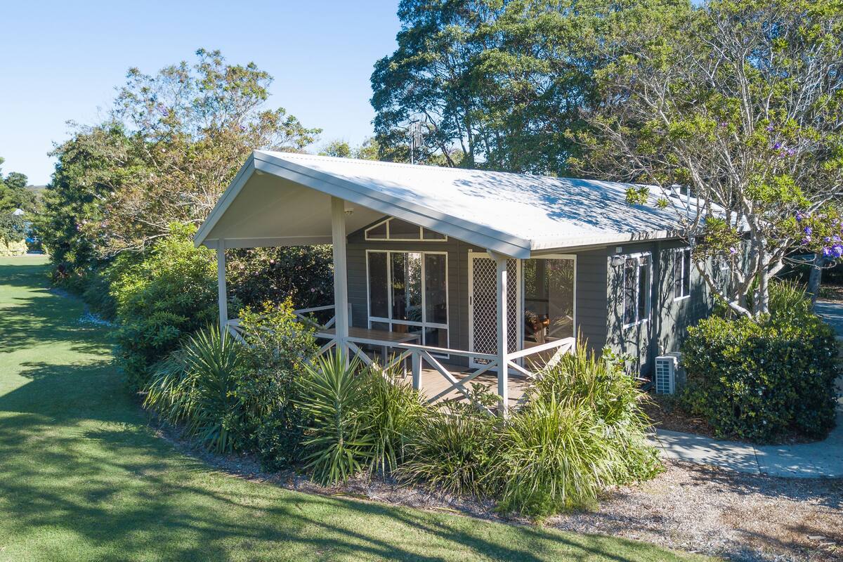 A small, single-story structure with a covered porch, surrounded by lush greenery and trees.
