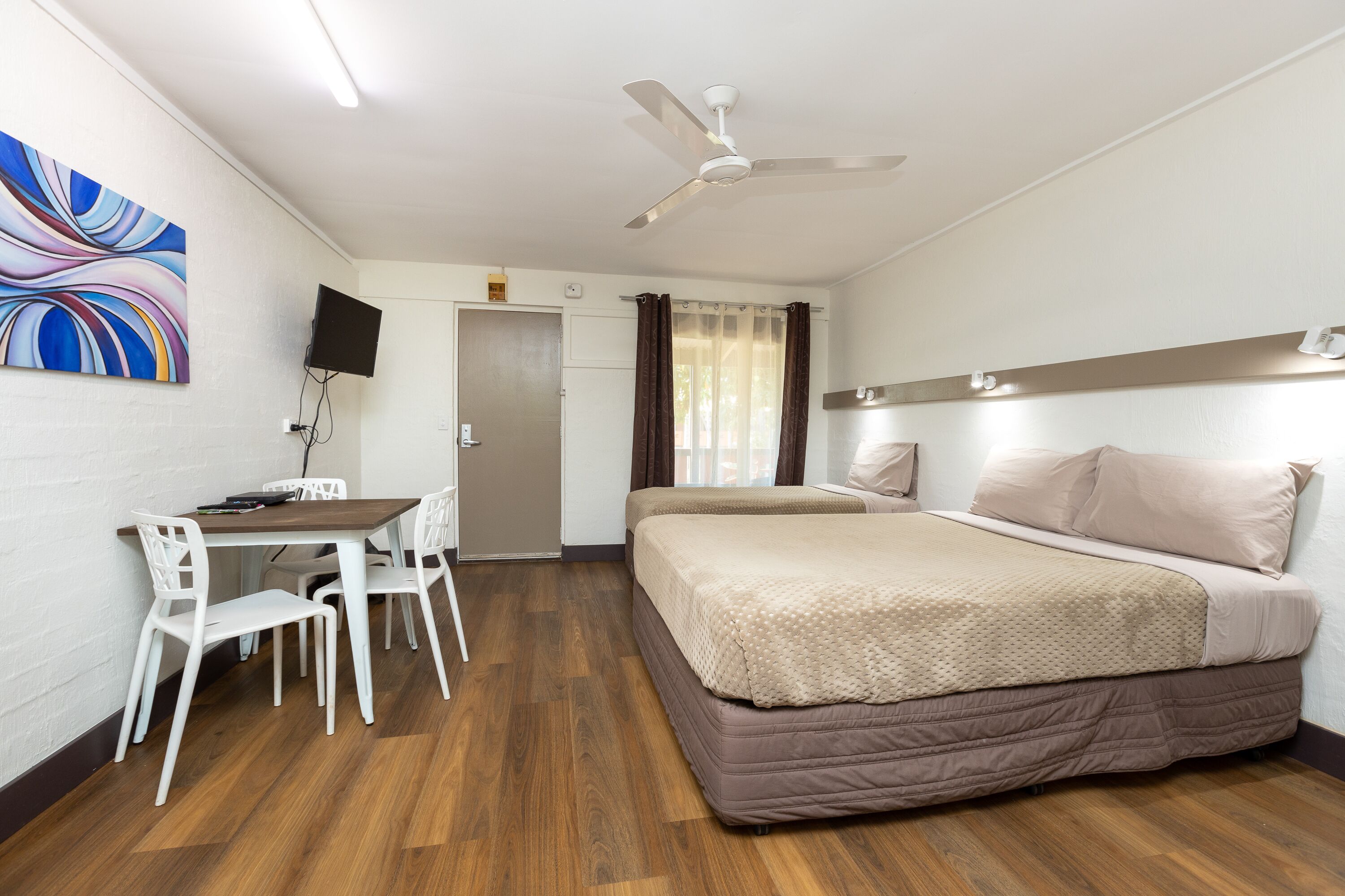 Una habitación de hotel con una cama, un escritorio con sillas, un ventilador de techo y un televisor colgado en la pared.