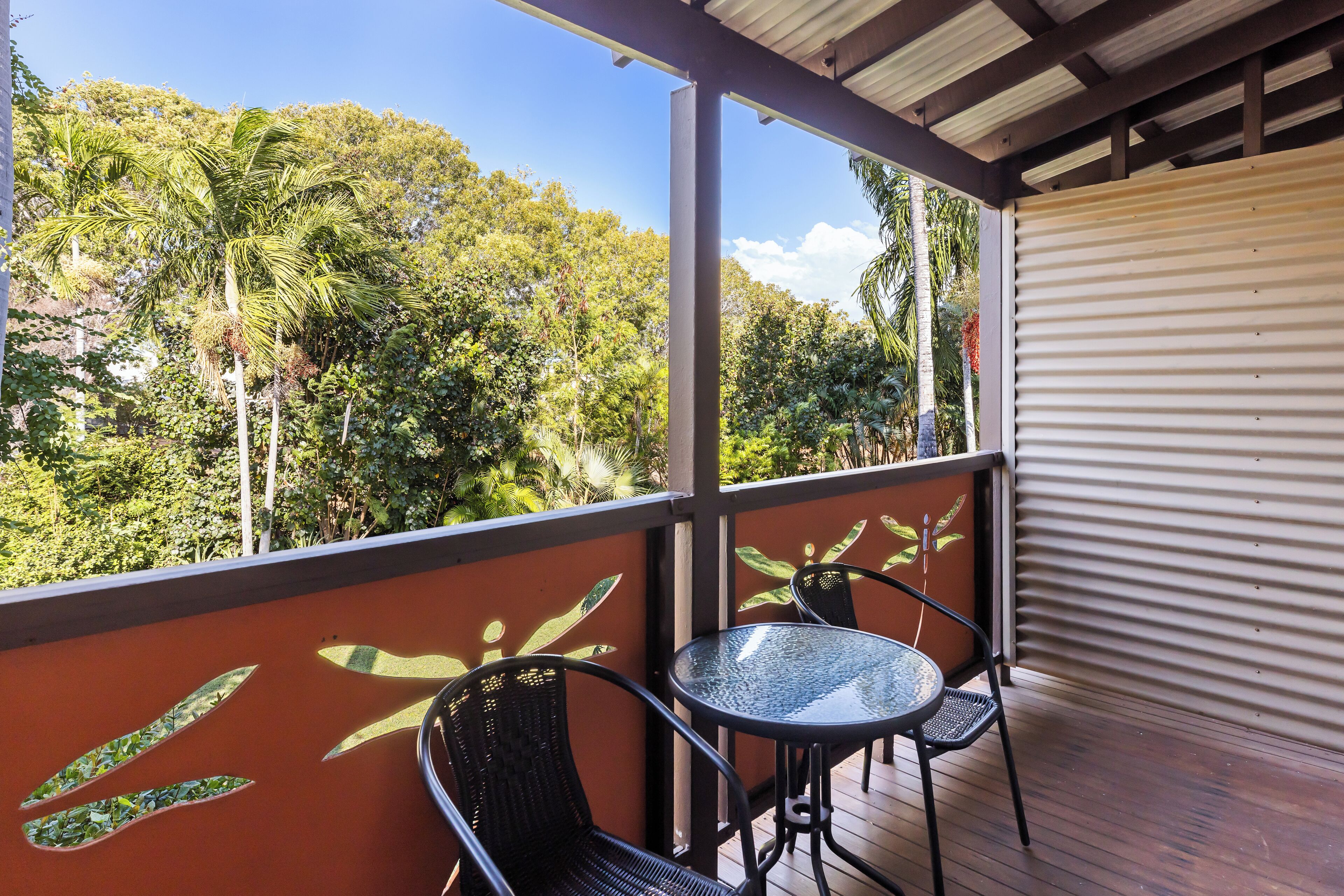 A covered outdoor area with two metal chairs and a glass table, overlooking a lush green landscape with trees and a clear sky.