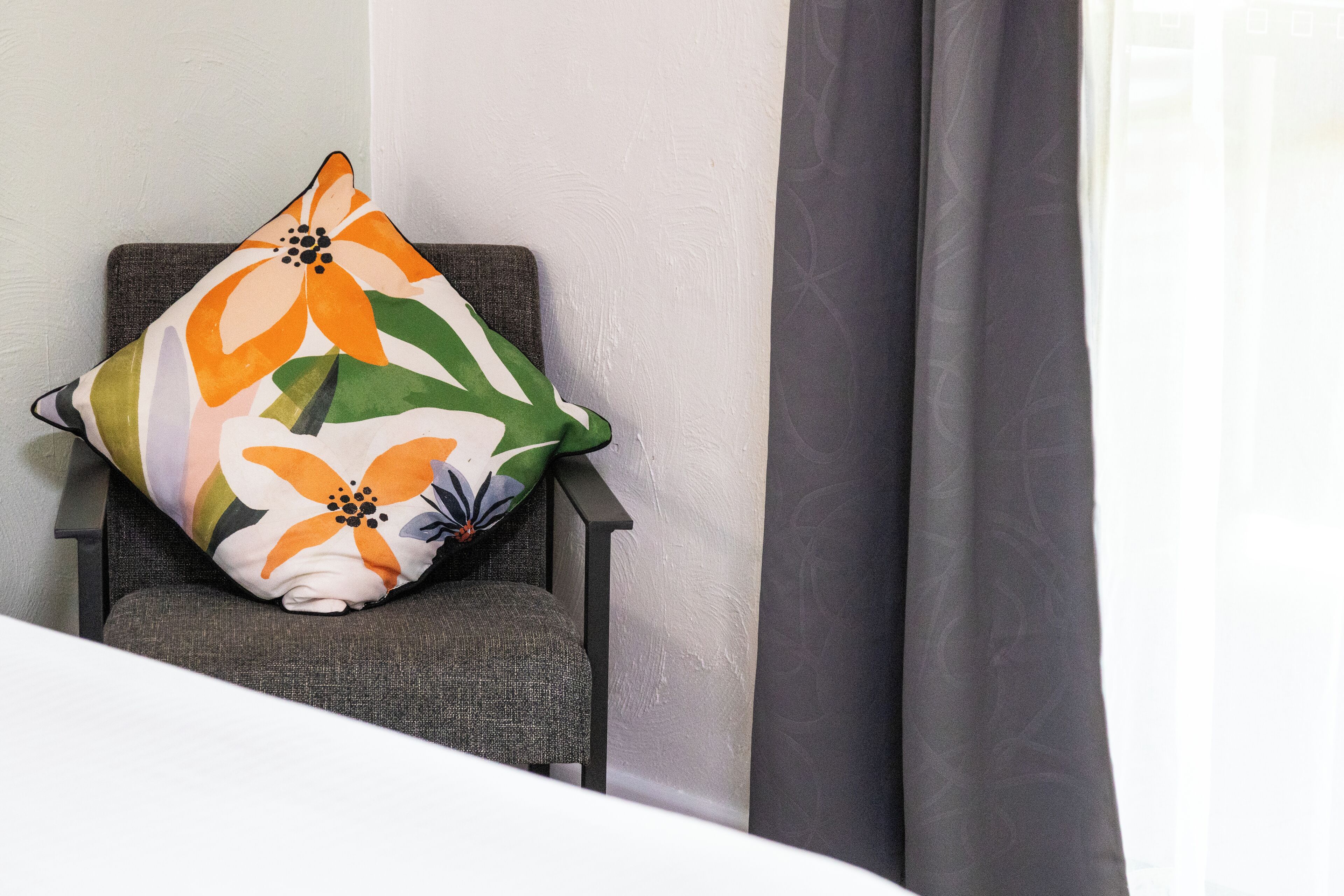 A chair with a floral pillow, a grey curtain, and a white wall.