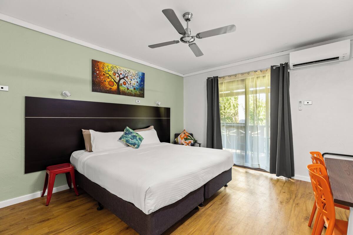 A bedroom with a bed, a ceiling fan, an air conditioning unit, a painting, and a view of the outdoors.