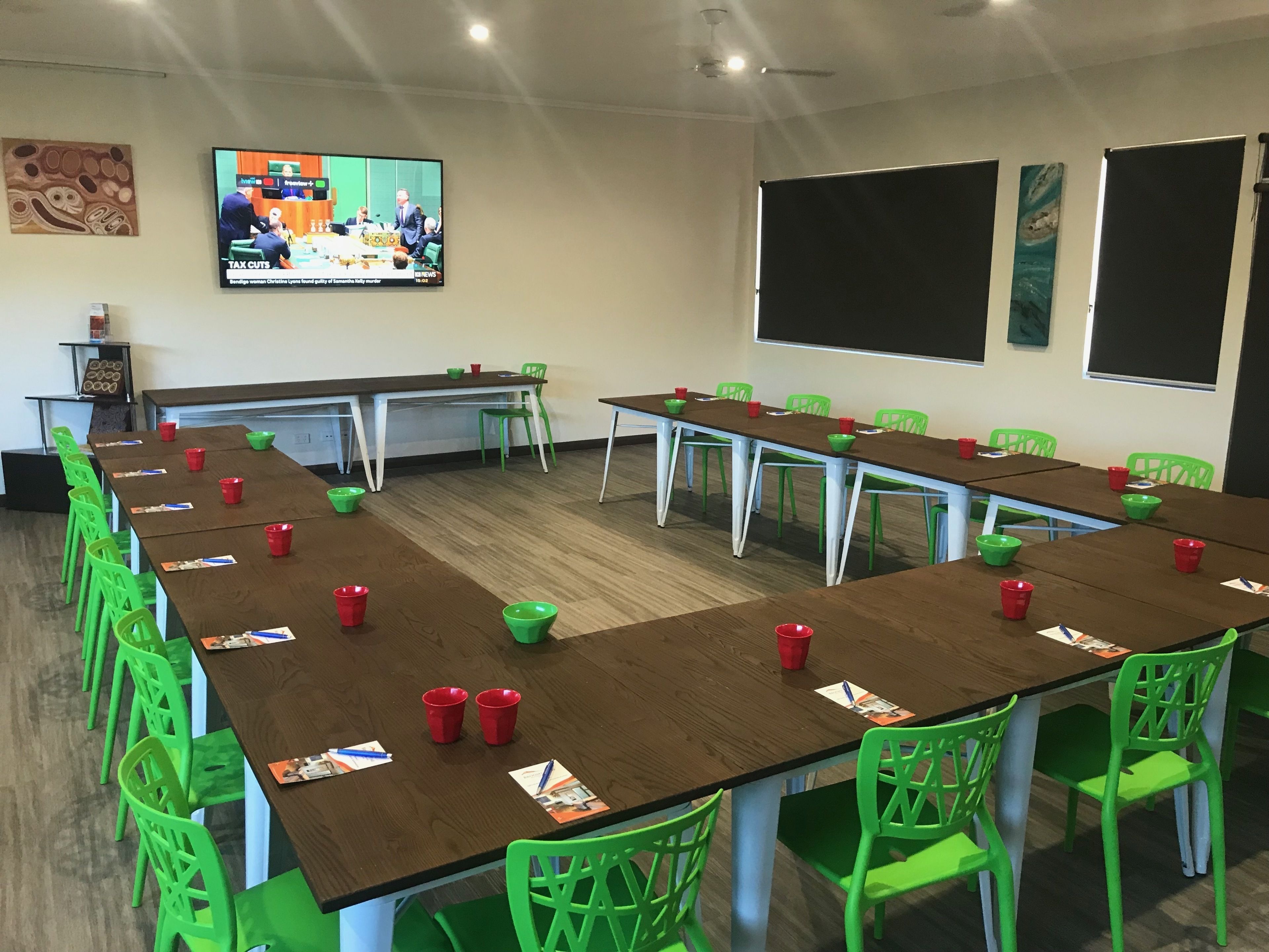 A conference room with multiple tables and chairs, a television screen displaying a news program, and a blackboard on the wall.
