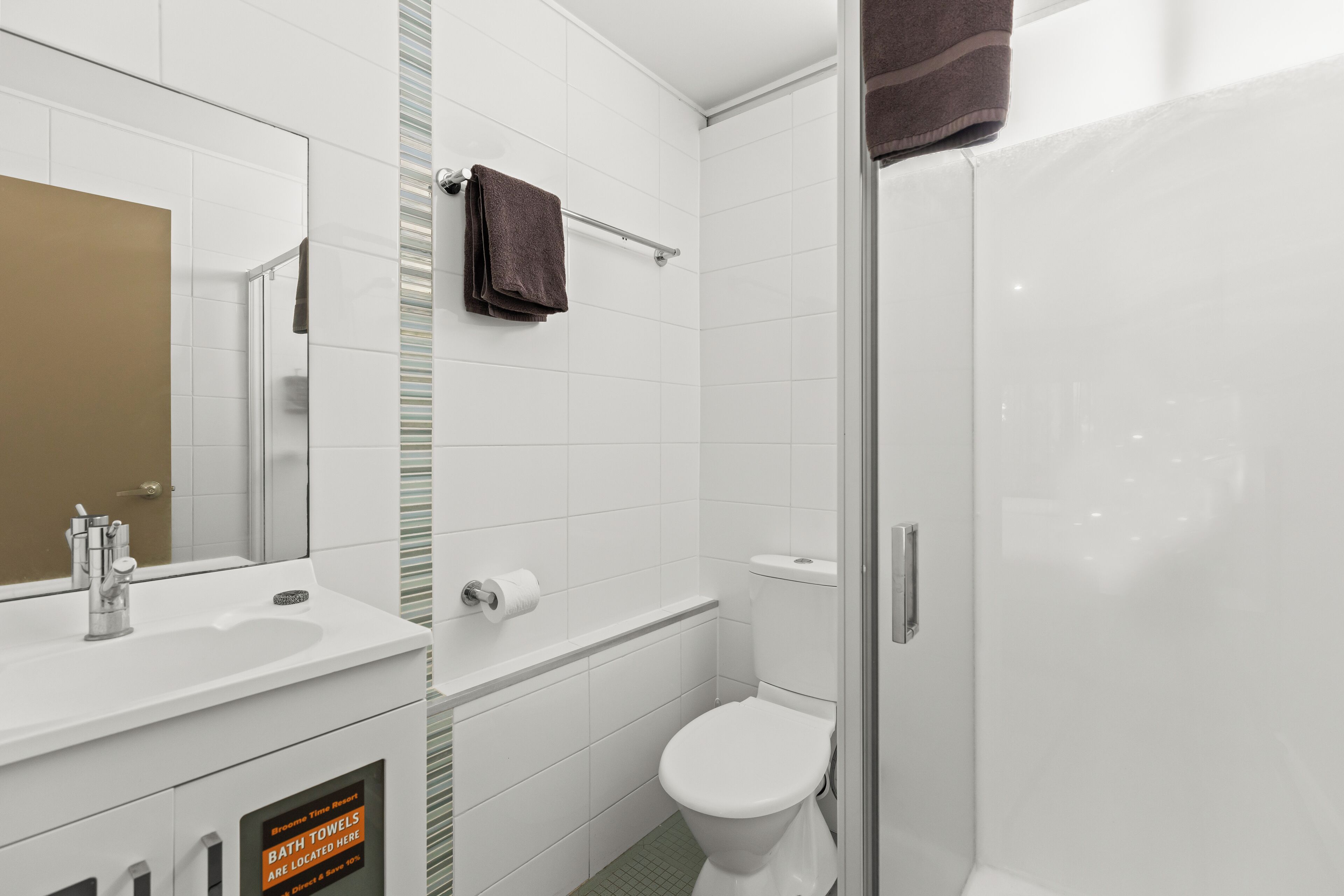 Ein Badezimmer mit weißem Waschbecken, Spiegel und Toilette, einem Handtuchhalter mit braunem Handtuch und einer Duschkabine aus Glas.