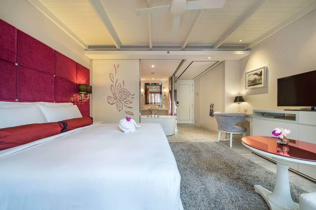 A hotel room with a large bed, a red headboard, a grey armchair, a small table with a vase, and a TV on the wall.