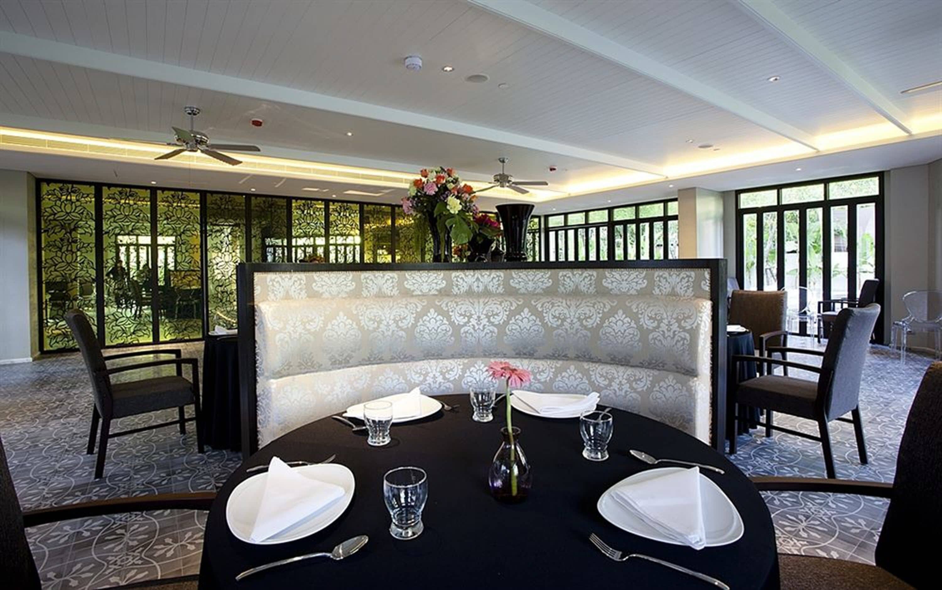 A dining area with a round table set for four, a bench with a patterned upholstery, and a view through a glass partition to an outdoor area with greenery.