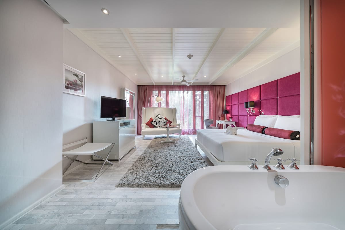 A modern hotel room with a large bed, a separate sitting area with a TV, a desk, and a bathroom with a bathtub and shower.