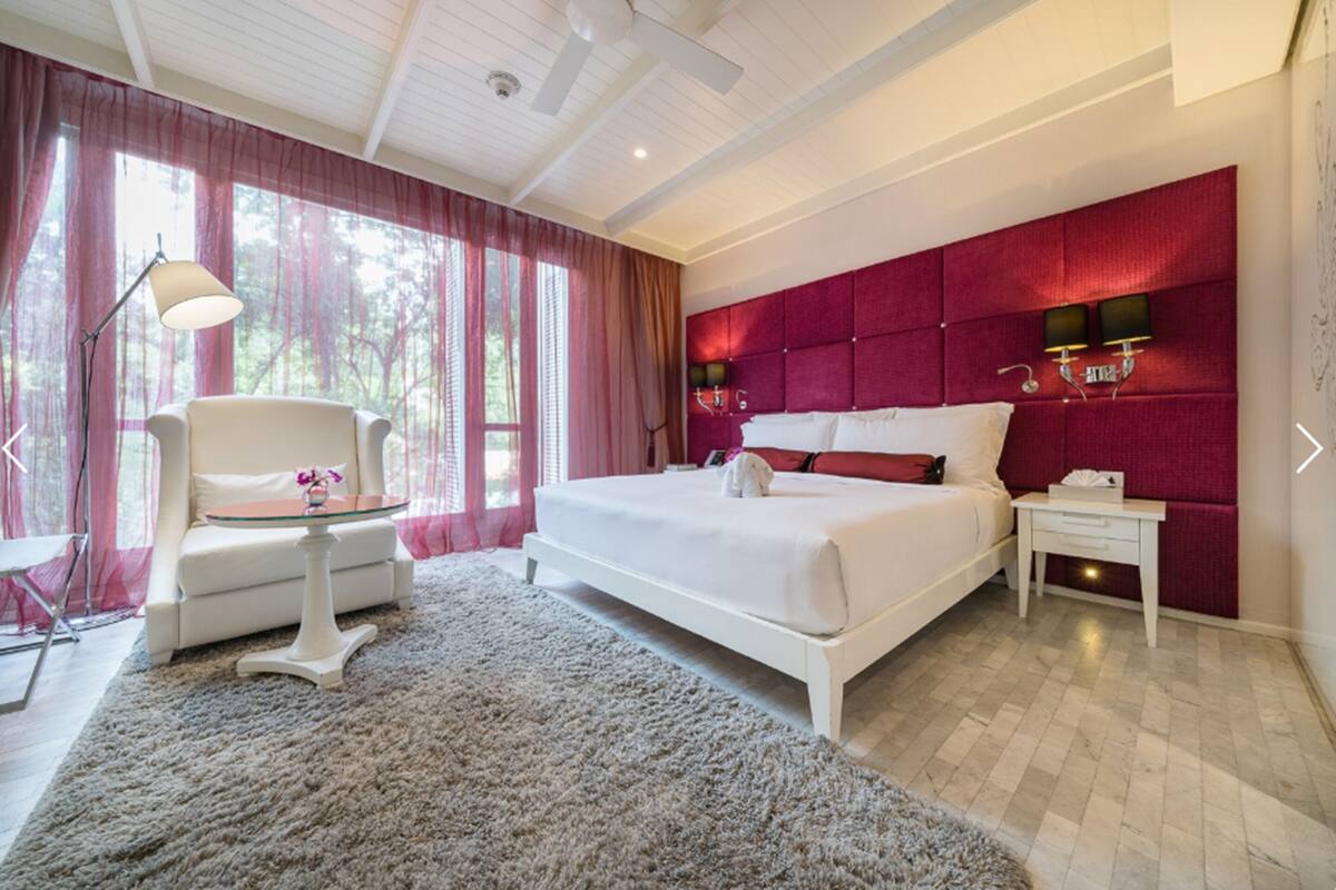 A modern bedroom with a large bed, a white armchair, a small table, and a red accent wall.