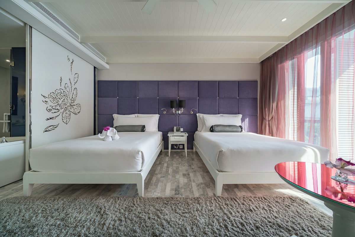 A modern bedroom with two beds, a glass coffee table, and a large window with sheer curtains.