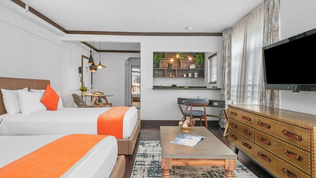 Quarto de hotel com duas camas, área de refeições, televisão e cómoda de madeira.
