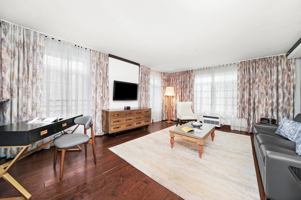 A modern living room with a flat-screen TV, a wooden desk, a grey chair, a sofa, and a coffee table.