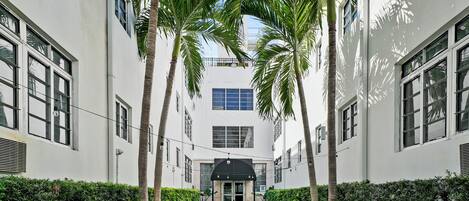 A narrow pathway flanked by white buildings and palm trees, leading to a central entrance with a black awning.