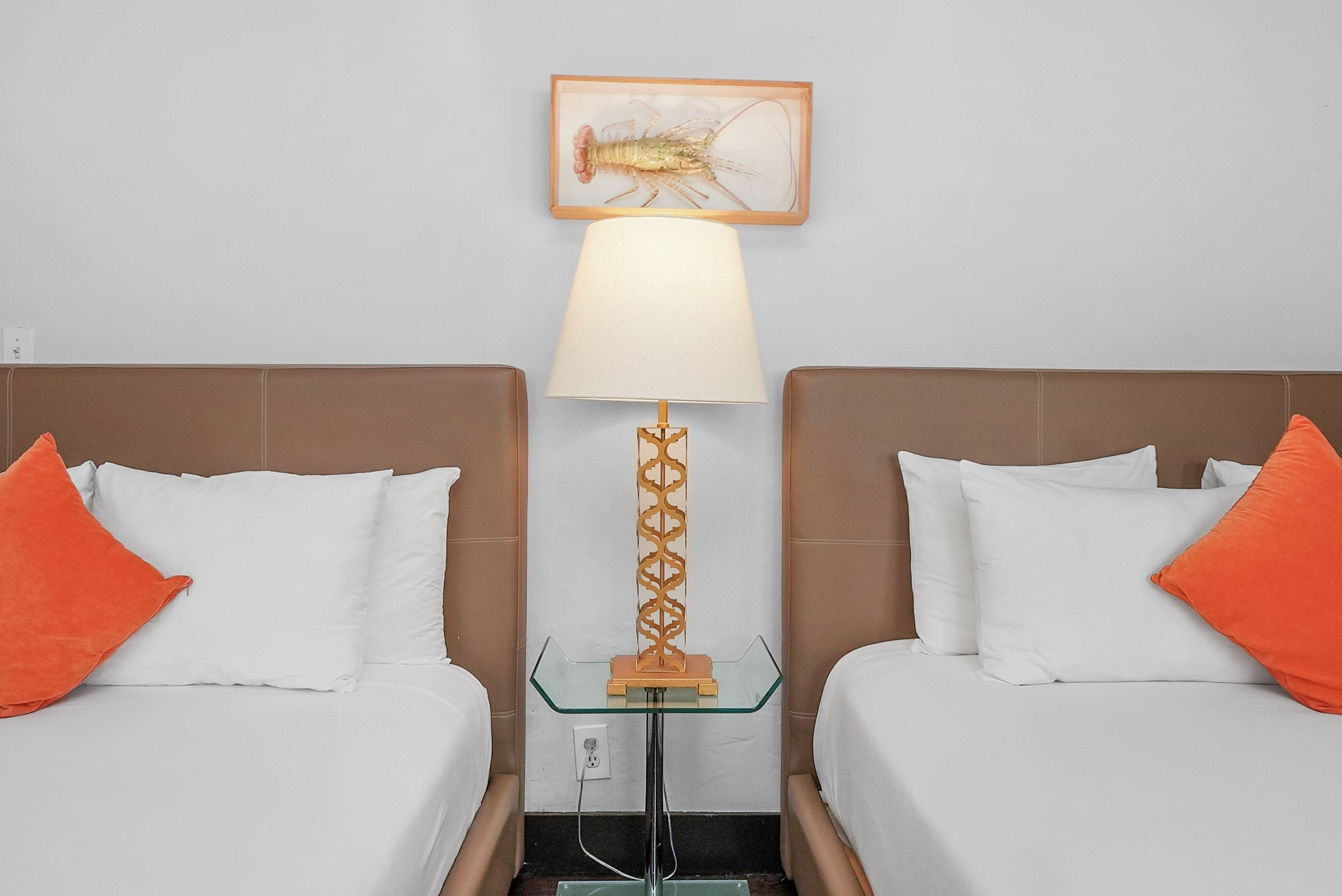 A hotel room with two beds, each with white bedding and orange pillows. A glass table with a wooden base is centered between the beds, and a lamp with a white shade is on the table. Above the table, there is a framed picture of a lobster on the wall.