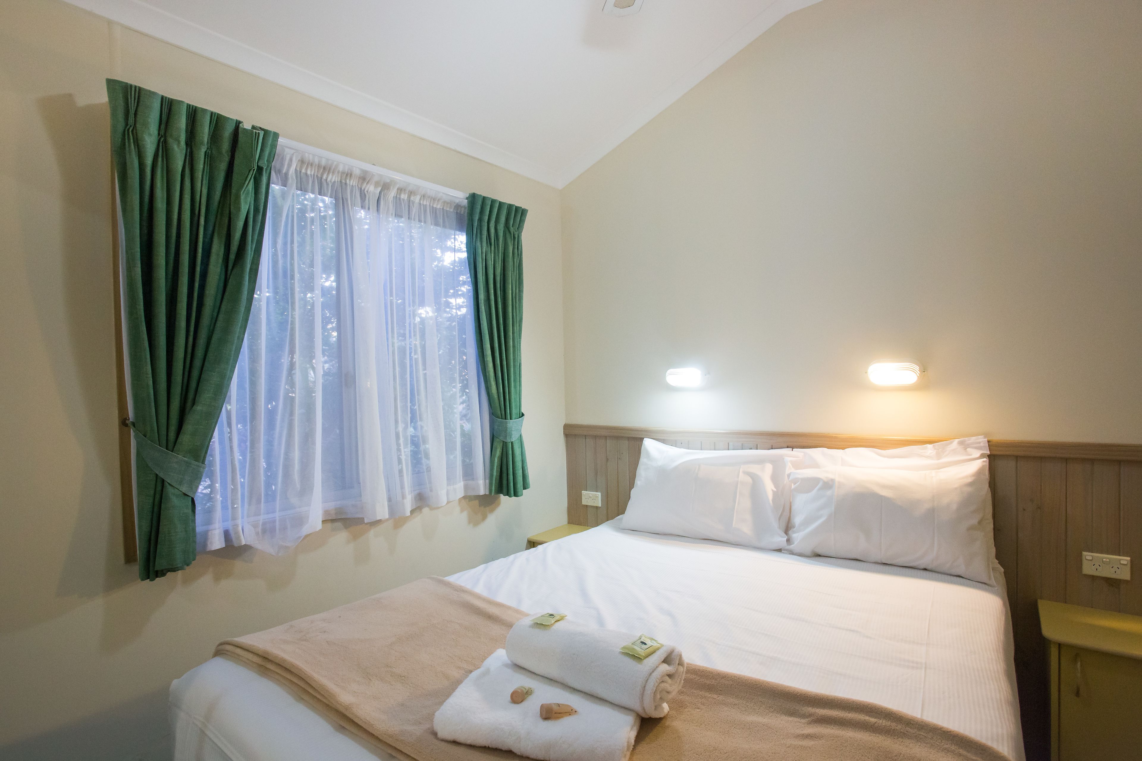 A hotel room with a bed, a nightstand, a window with curtains, and a wall-mounted light.