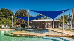 Una piscina cristalina con una zona sombreada, sillas de descanso y un edificio con un letrero.