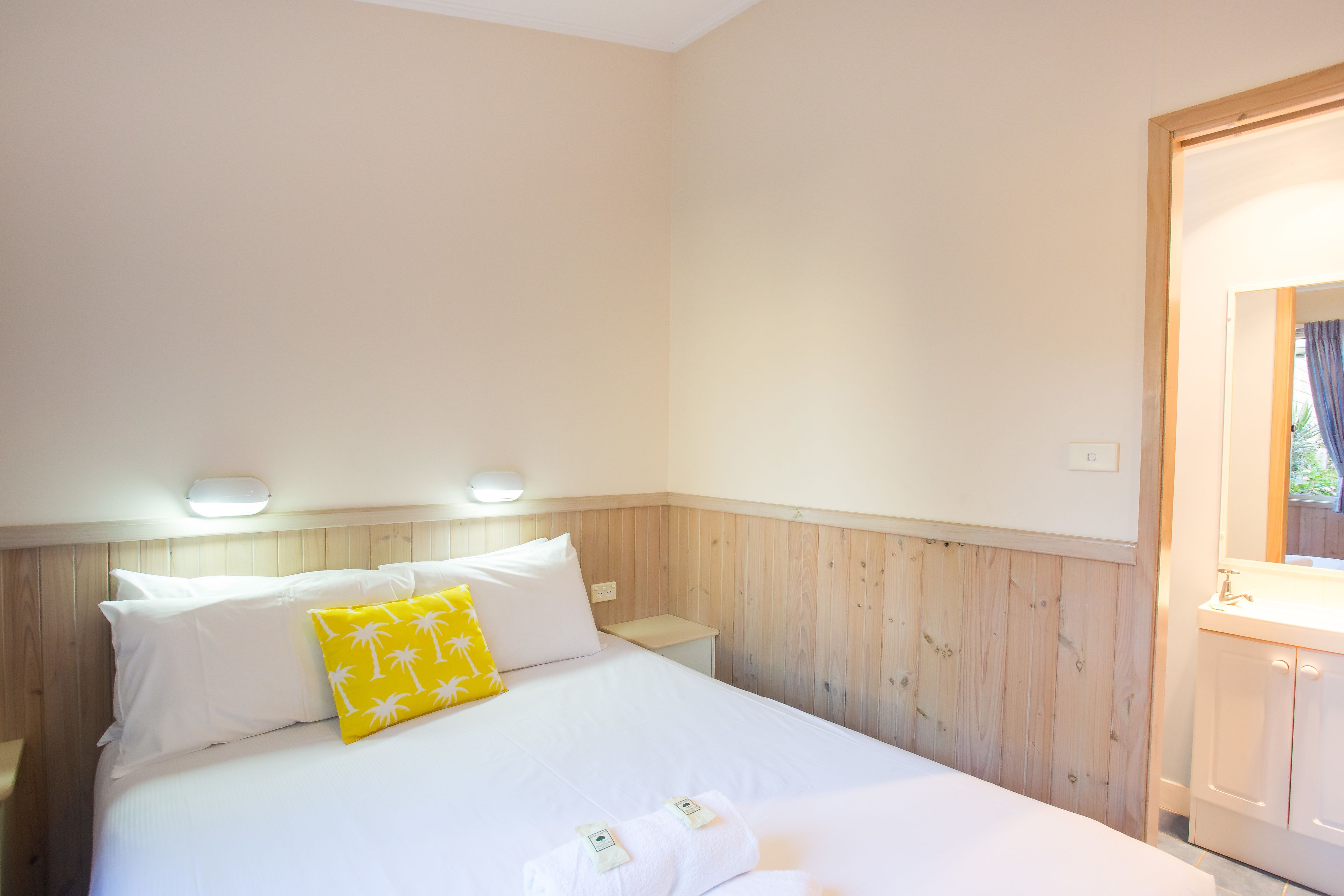 A neatly made bed with white linens and a single yellow pillow, a wooden headboard, and a small bedside table with a lamp.