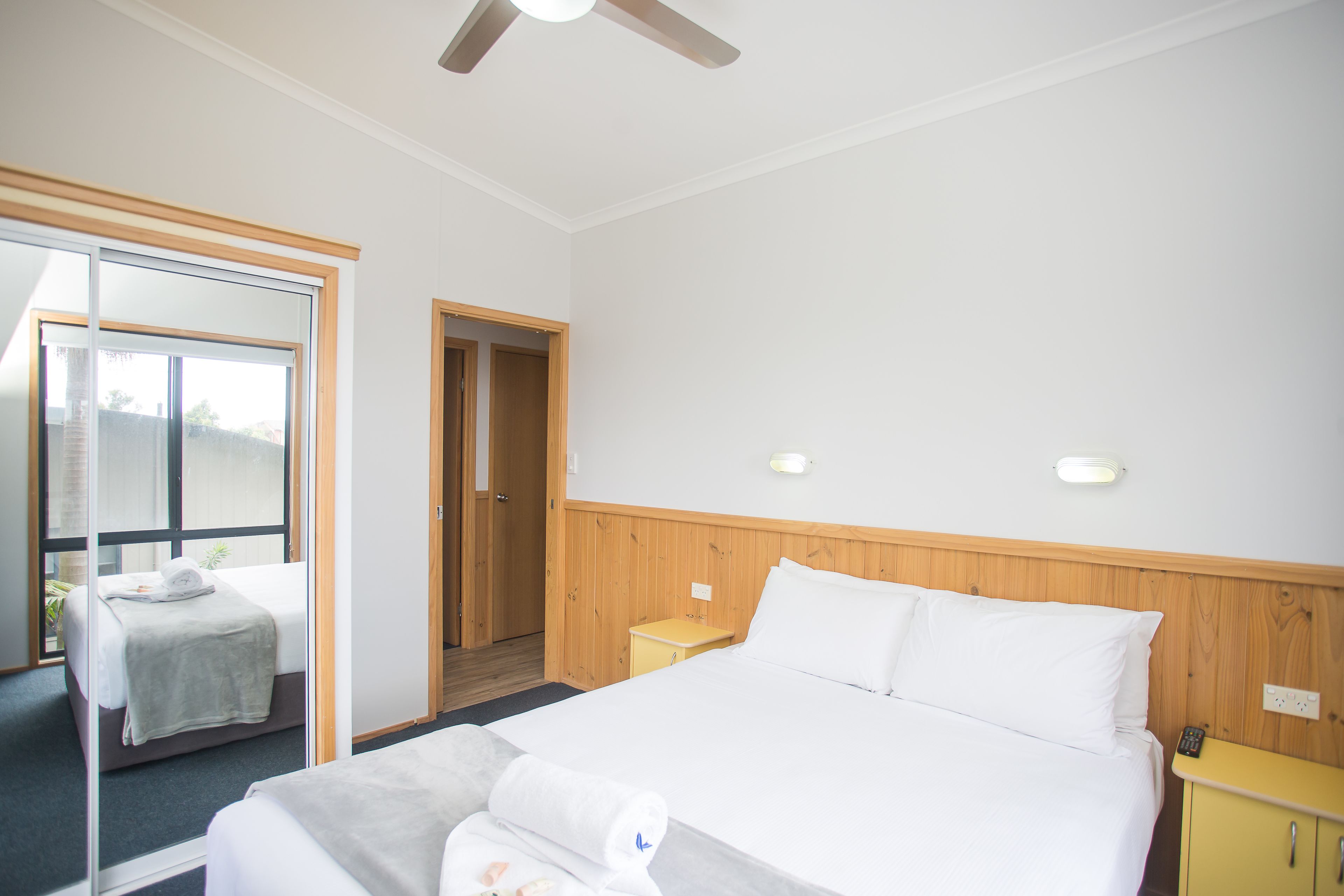Ein Hotelzimmer mit einem großen Bett, einem hölzernen Kopfteil, einem Nachttisch, einem kleinen gelben Schrank und einer Schiebetür zu einem weiteren Raum.