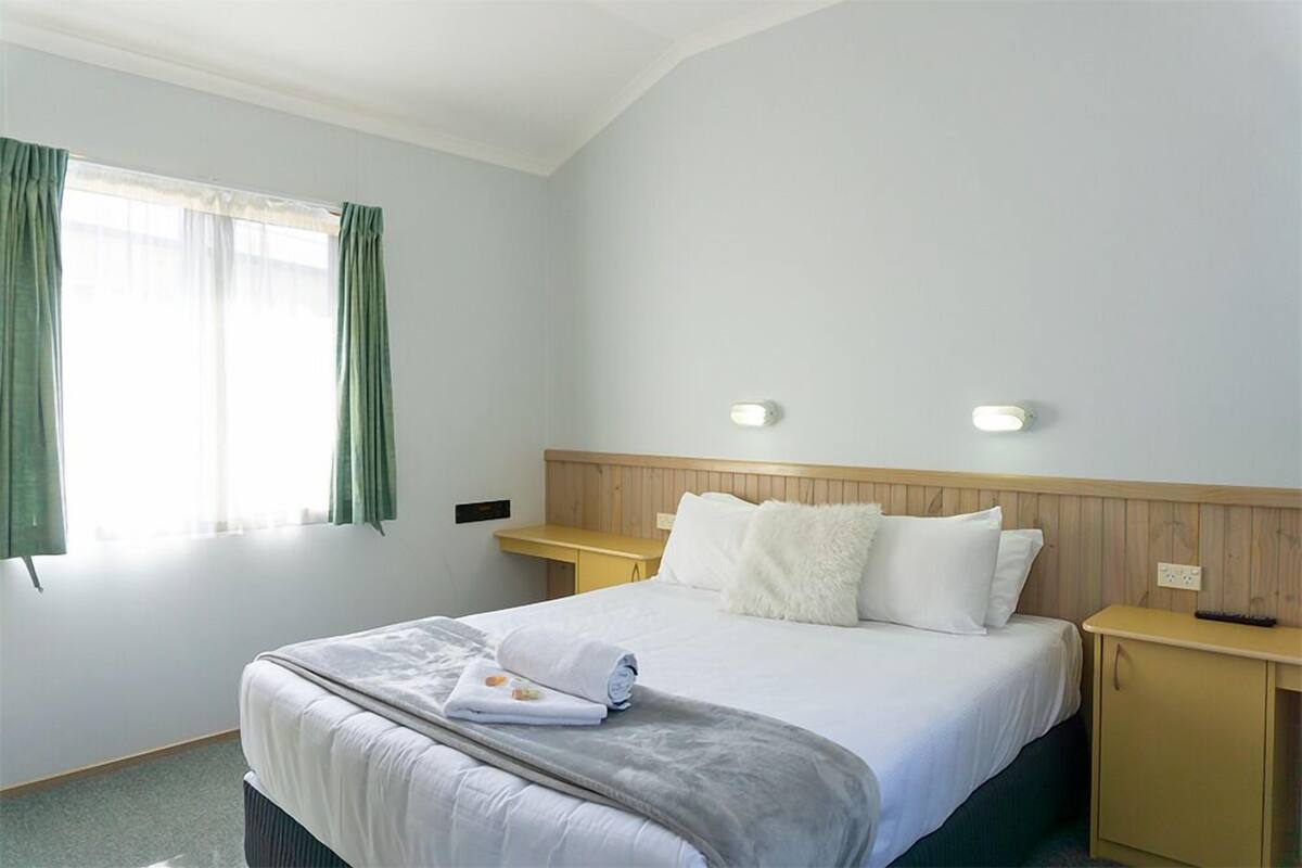 Ein Hotelzimmer mit einem ordentlich bezogenen Bett, einem hölzernen Kopfteil, zwei Nachttischen und einem Fenster mit Vorhängen.
