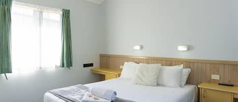 A hotel room with a neatly made bed, a wooden headboard, two bedside tables, and a window with curtains.