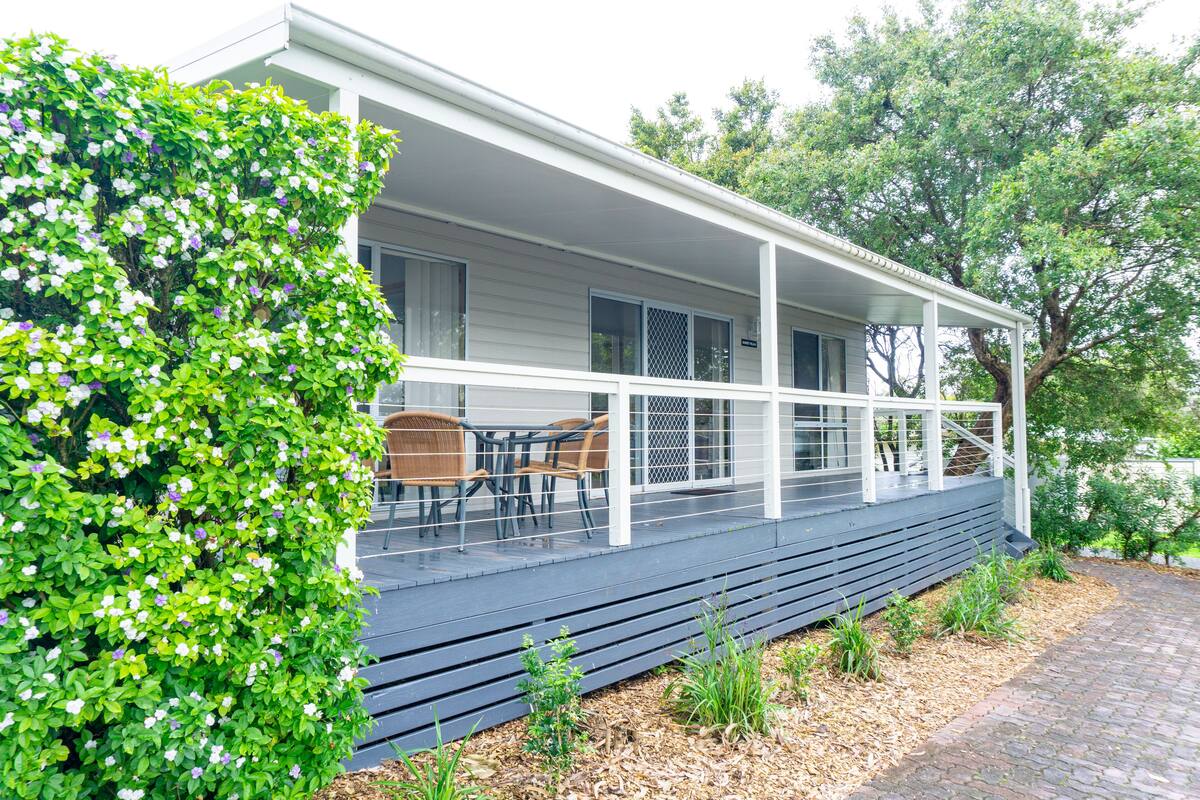 Ein Haus mit überdachter Veranda, einem Essbereich mit Stühlen und einem kleinen Garten mit Pflanzen und Mulch.
