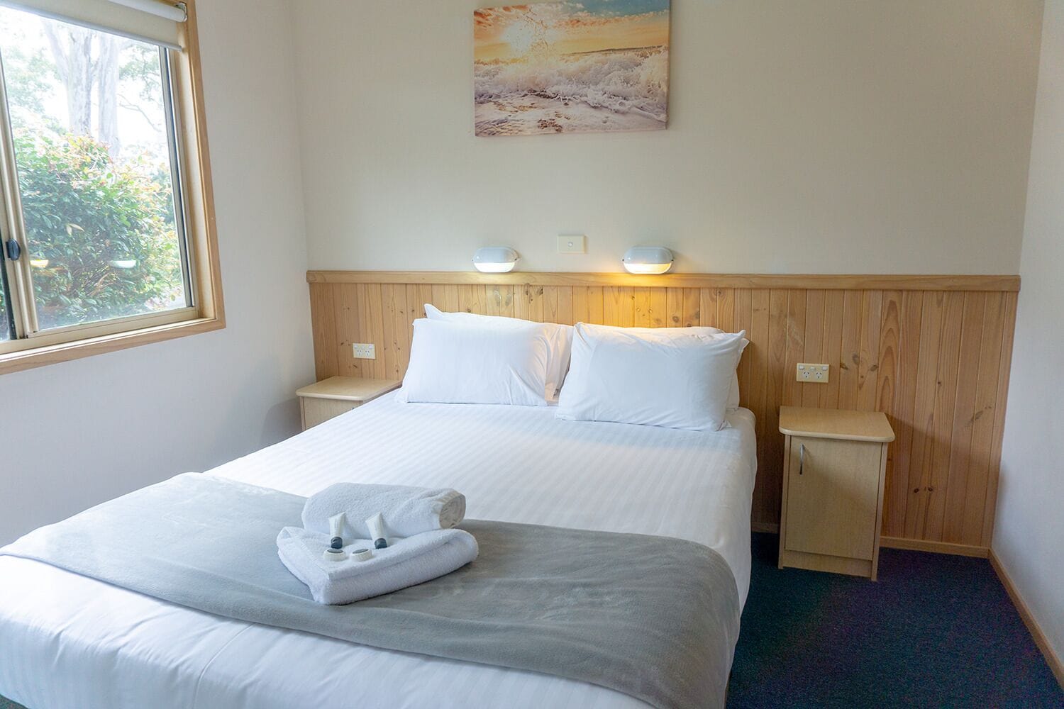 A neatly made bed with white linens, two bedside tables with lamps, a painting on the wall, and a window with a view of greenery.