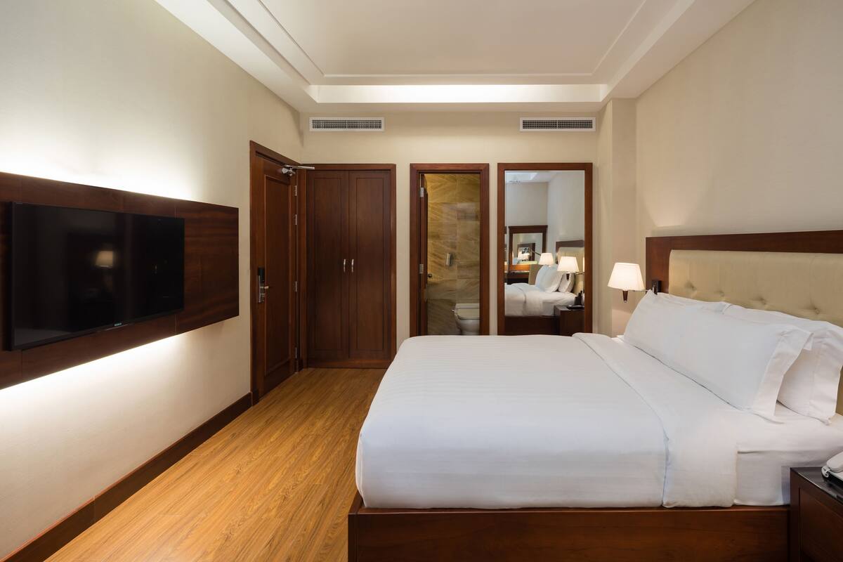 A hotel room with a large bed, wooden flooring, a flat-screen TV mounted on the wall, a wardrobe, and a bathroom visible through an open door.