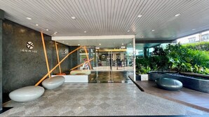 Una entrada de edificio moderna con fachada de vidrio, una rampa que conduce a la entrada y un área de descanso con bancos.
