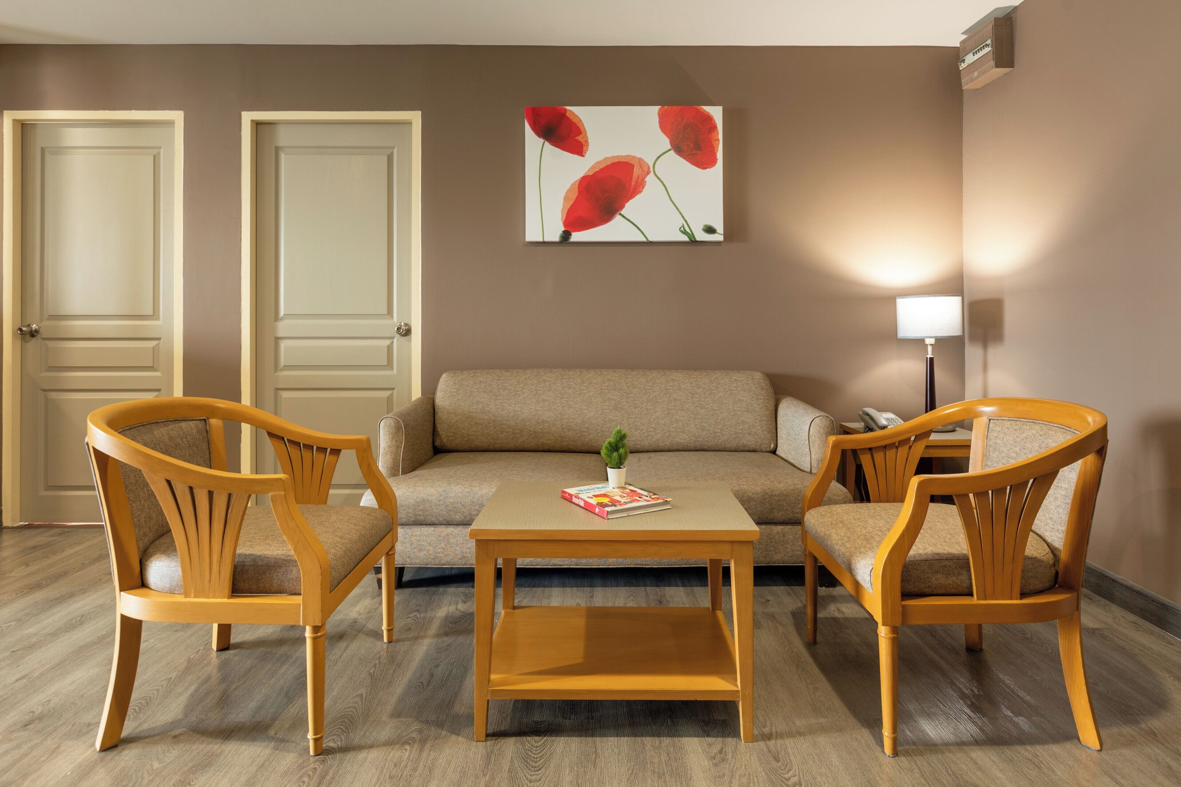 A room with a sofa, two wooden chairs, a coffee table, and a painting of red flowers on the wall.