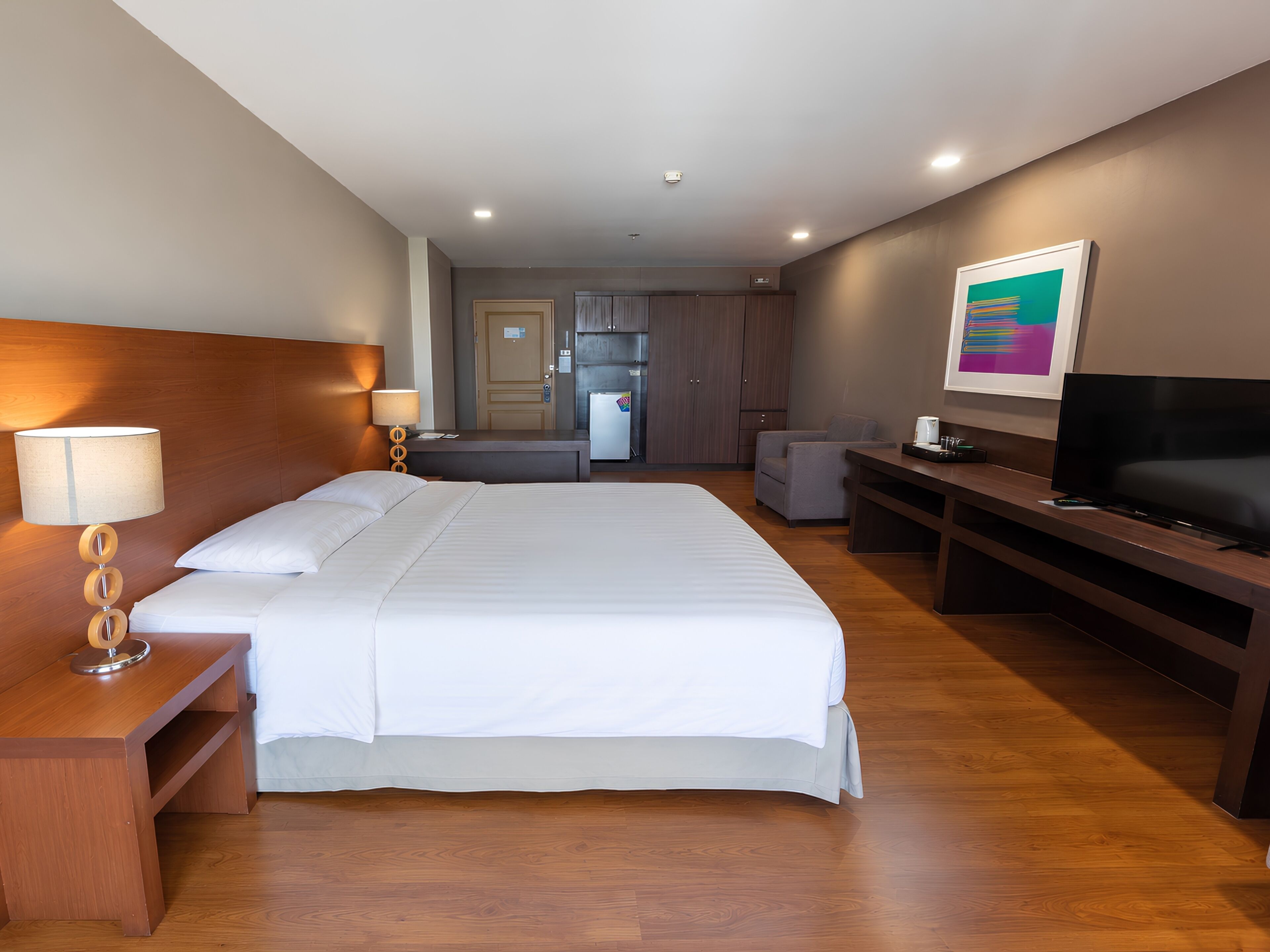 A hotel room with a large bed, bedside tables, a TV, a desk, and a kitchenette in the background.
