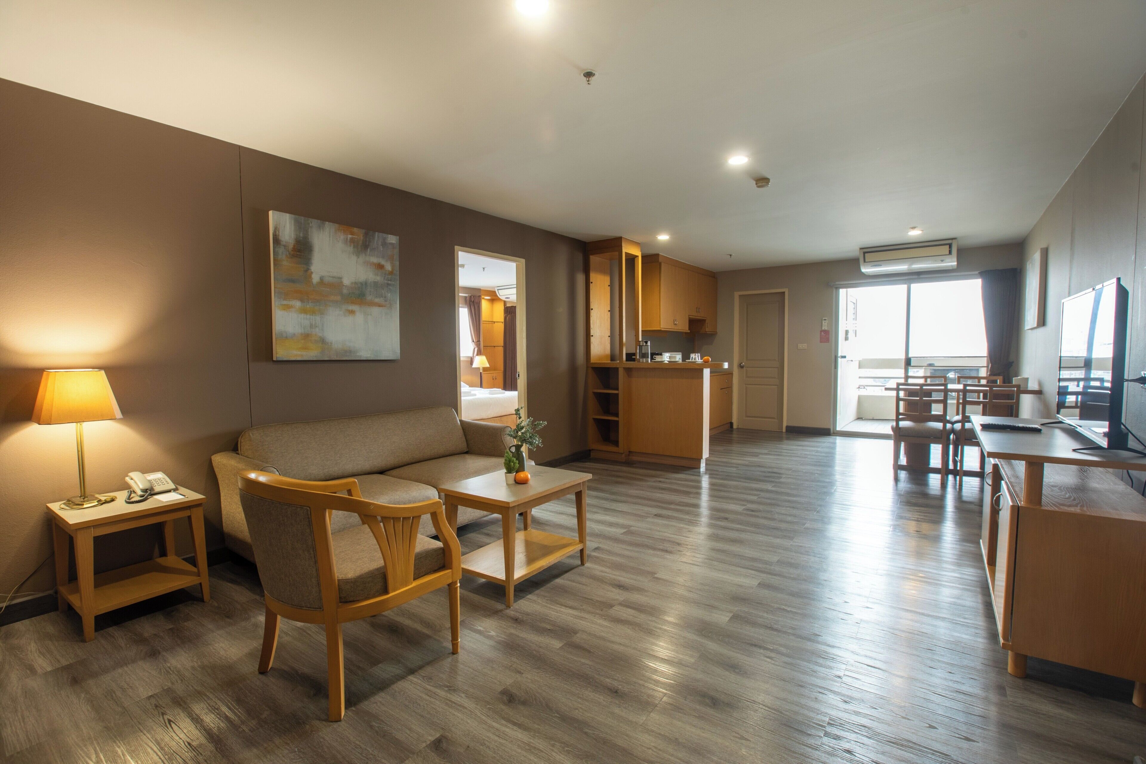 Une chambre d’hôtel moderne équipée d’un canapé, d’un fauteuil, d’une petite table et d’un bureau avec une chaise. Elle comprend également un coin cuisine avec des placards et un four à micro-ondes. La chambre est dotée d’un plancher en bois et d’une grande fenêtre qui laisse entrer la lumière naturelle.