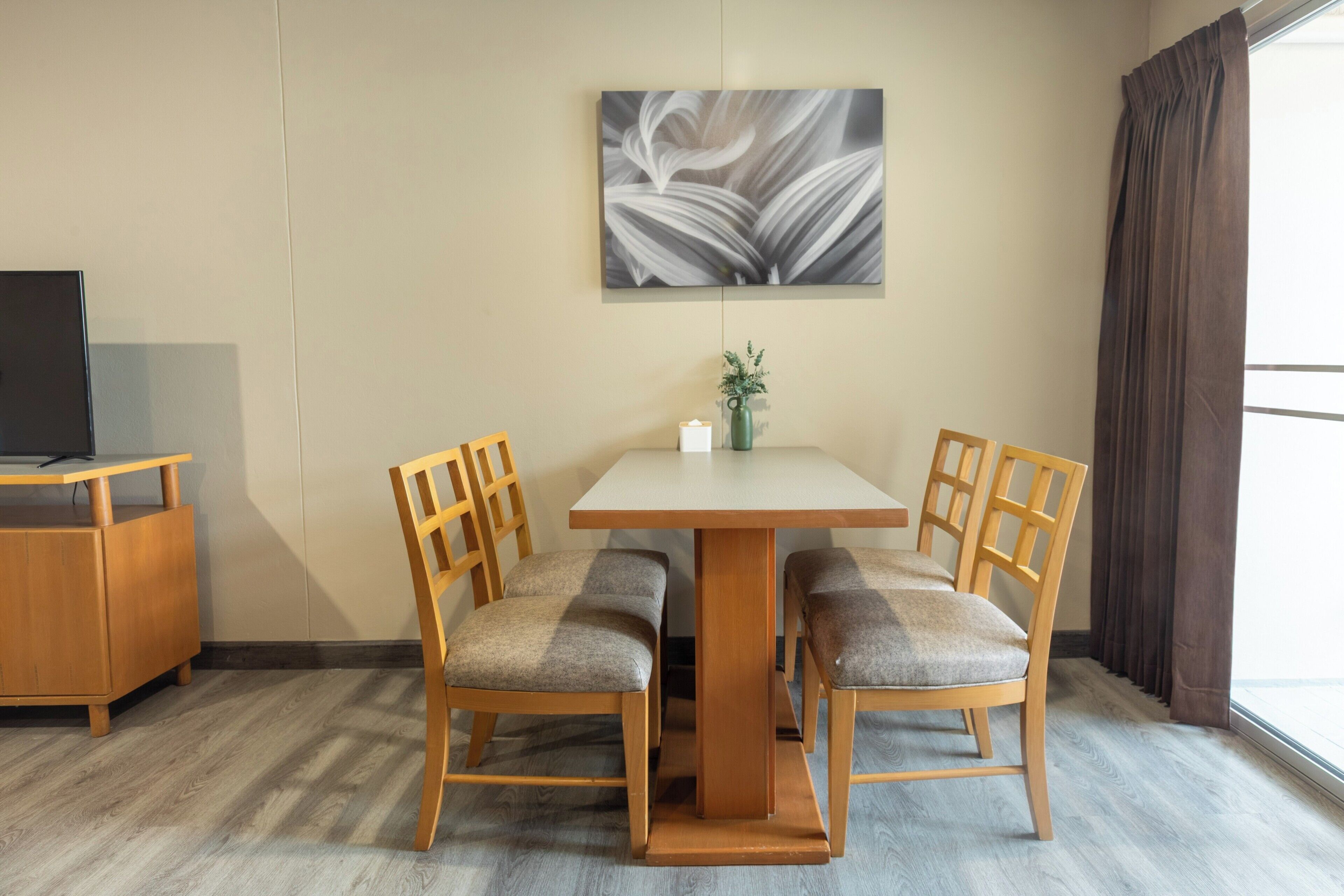 A hotel room with a dining table, four chairs, a television on a stand, a painting on the wall, and a window with curtains.