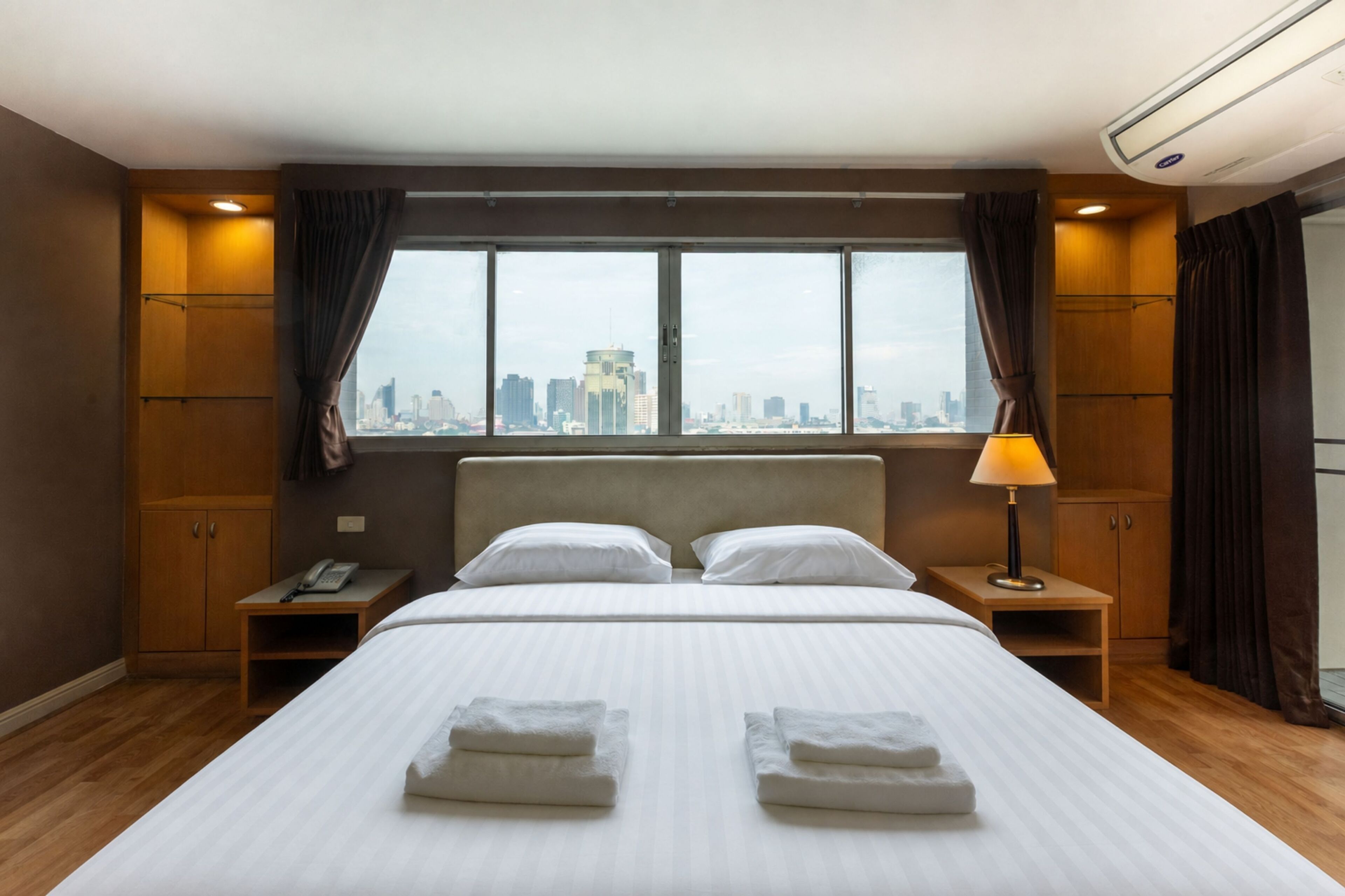 A hotel room with a large bed, two bedside tables, a lamp, a phone, and a city view through the window.
