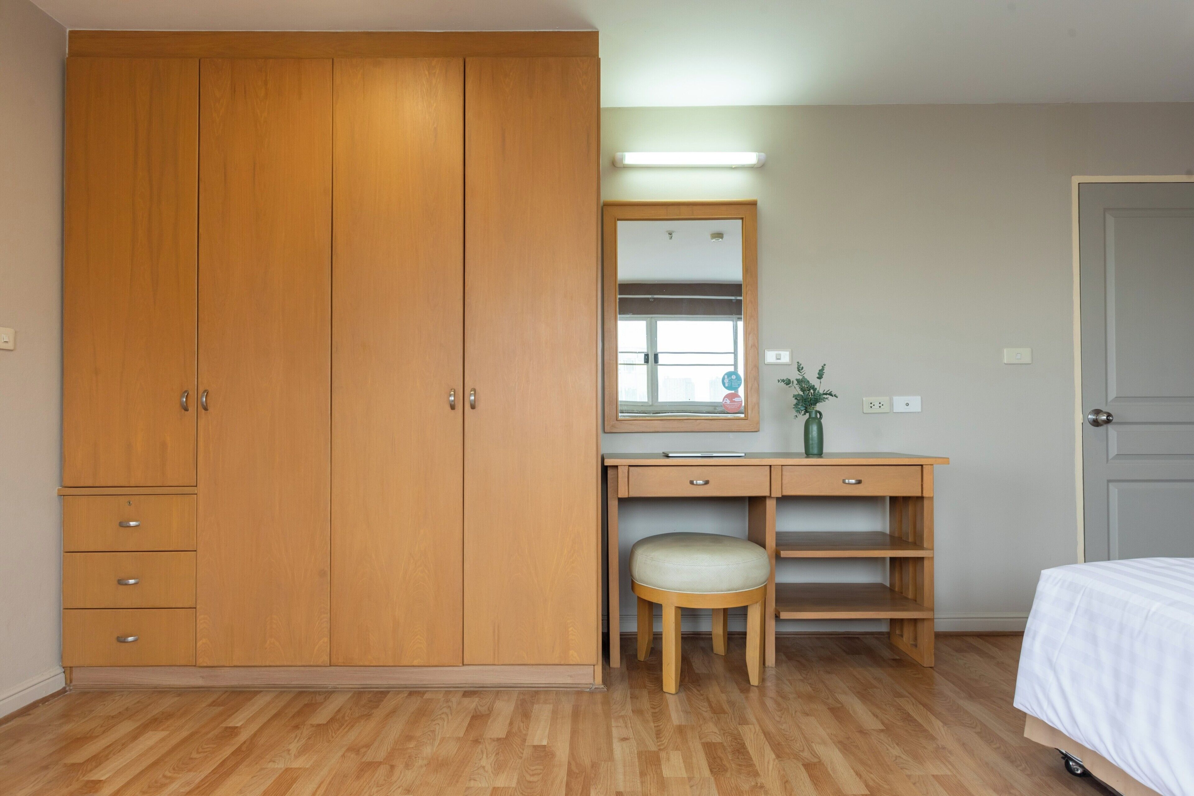 A bedroom with a wooden wardrobe, a desk with a mirror, a chair, and a bed.