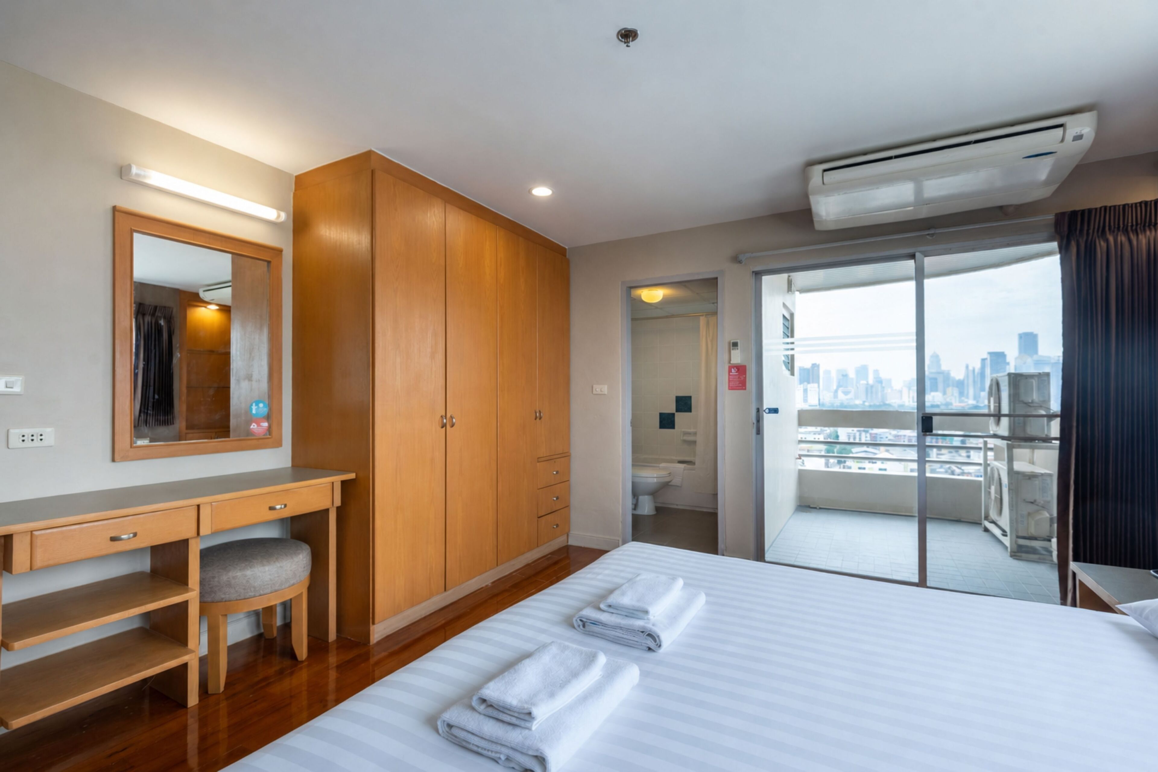 A hotel room with a bed, a desk, a mirror, a wardrobe, and a view of the city.